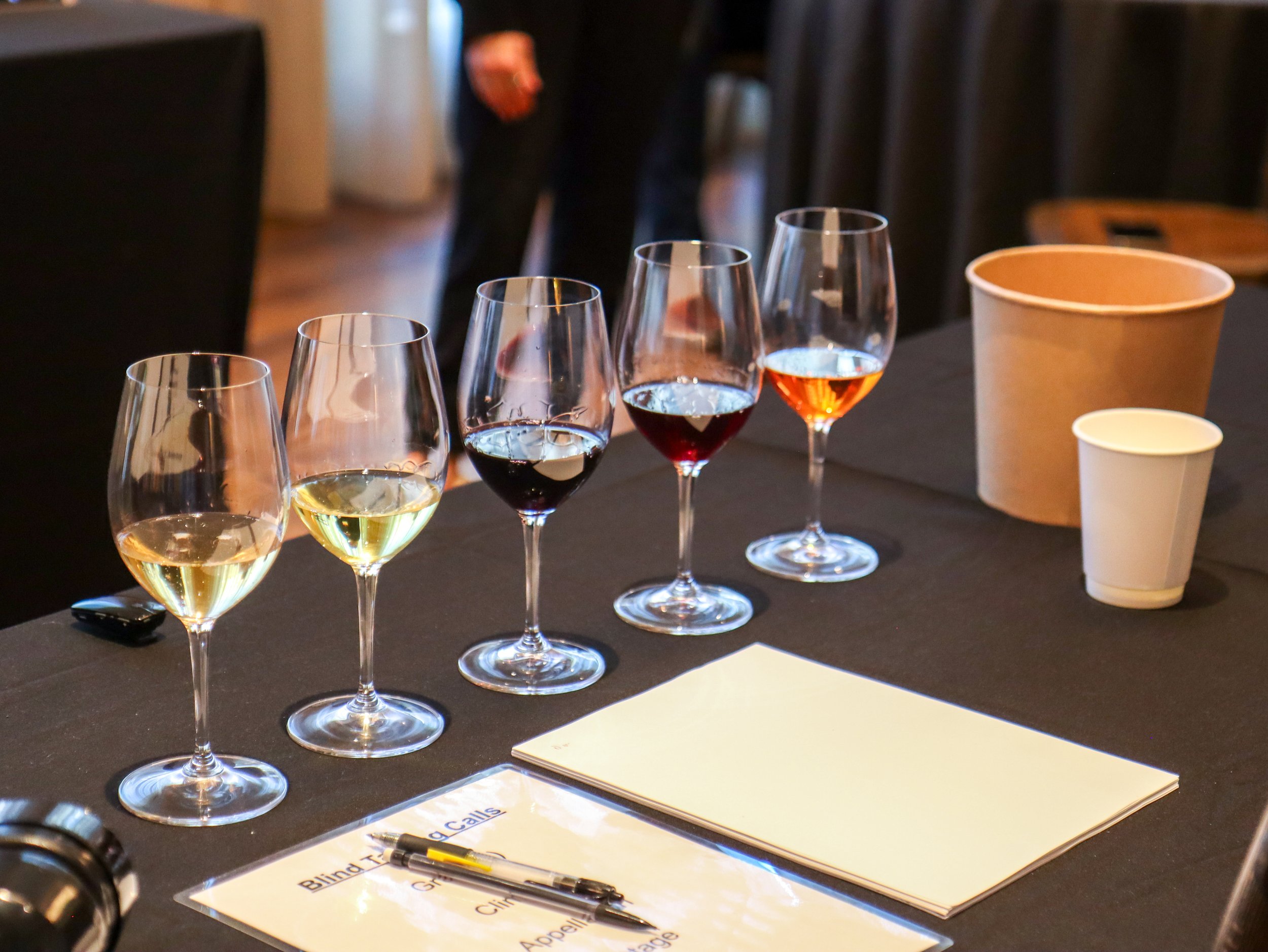Six wine glasses filled with white wine, red wine, and rosé on a dark table. There is a paper sheet, pens, a disposable coffee cup, and a partially visible clause sheet in the foreground. A person in dark clothing stands in the background.