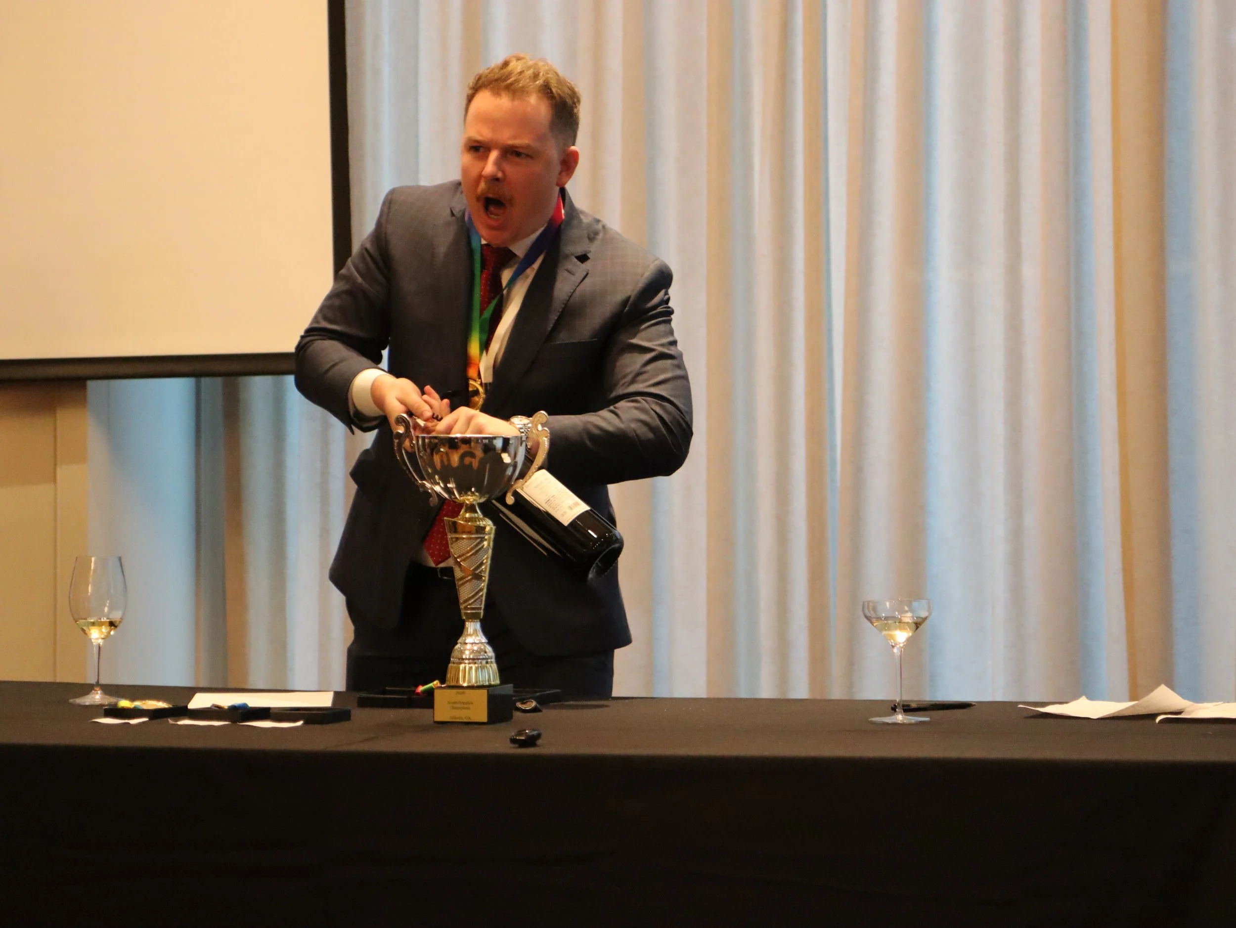 A man in a suit has an angry expression while holding a bottle of wine at a table with a trophy and glasses, in front of a curtained wall.