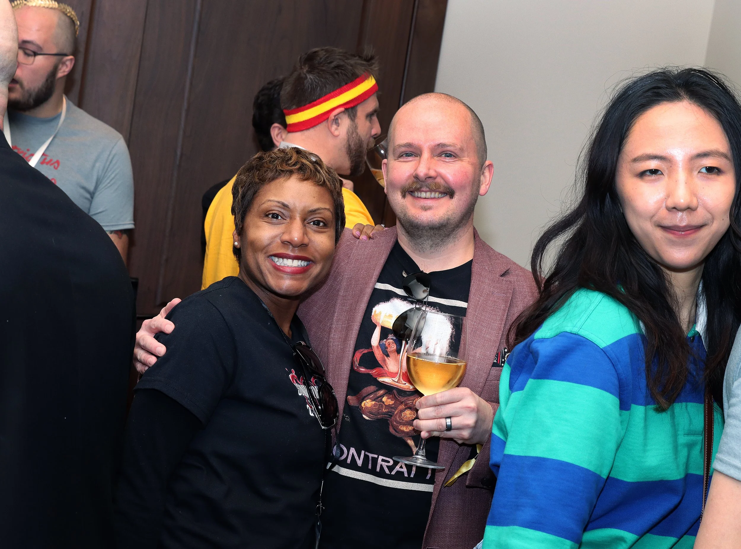 A group of friends celebrating at a party, smiling and holding drinks, with one person wearing a red and yellow headband.