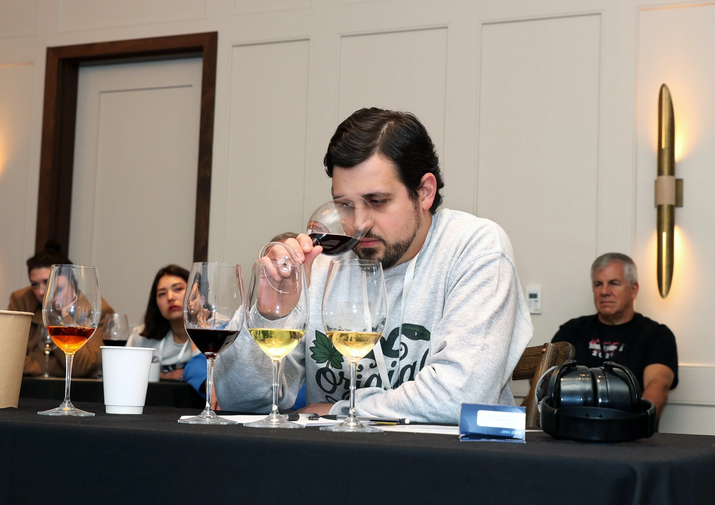 A man with dark hair and beard is seated at a table, holding a glass of red wine close to his nose, seemingly smelling it, with a focused expression. In front of him are four wine glasses with different types of wine, and other people are sitting aro