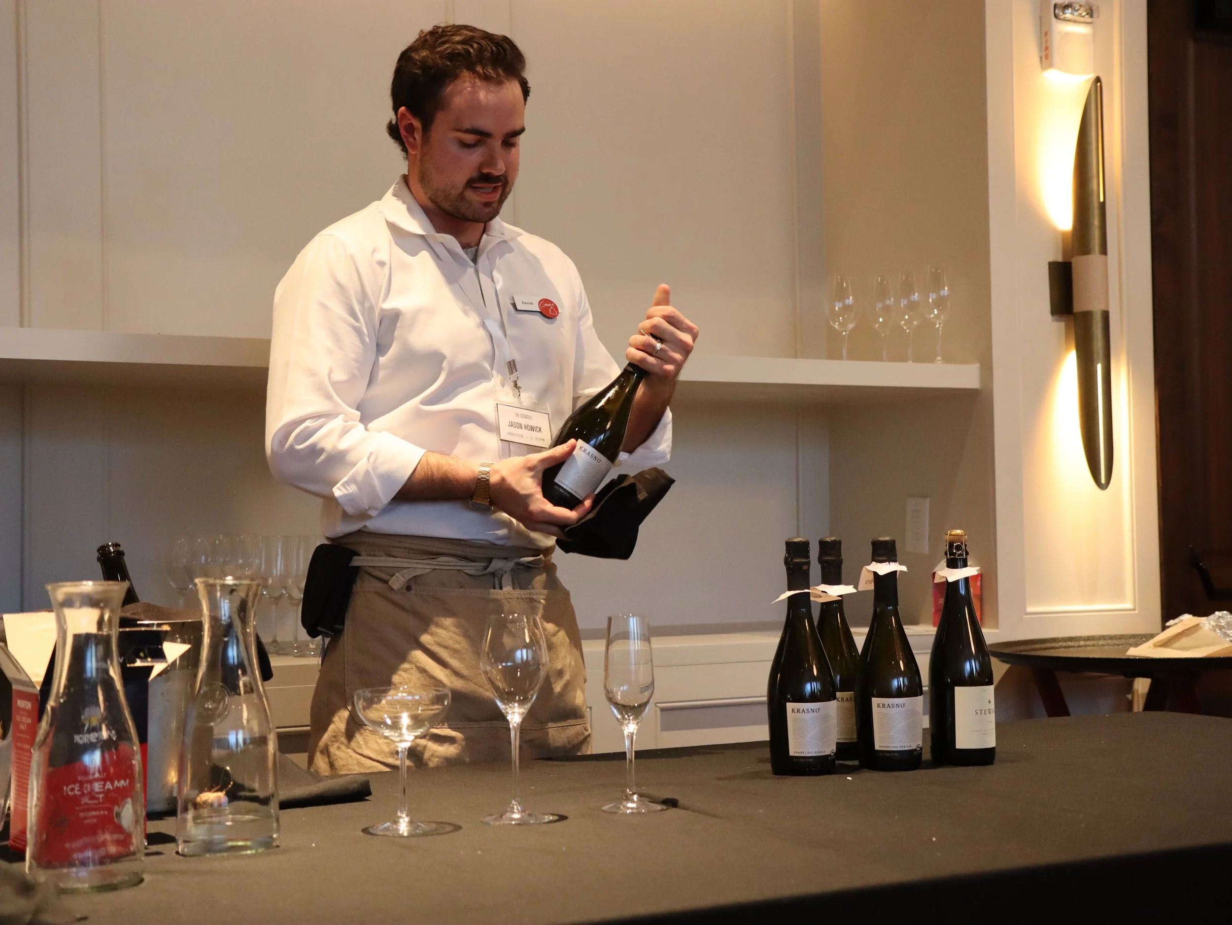 A man with a name tag reading Jason Howick holding a wine bottle at a tasting event, surrounded by glasses and bottles of sparkling wine on a table.