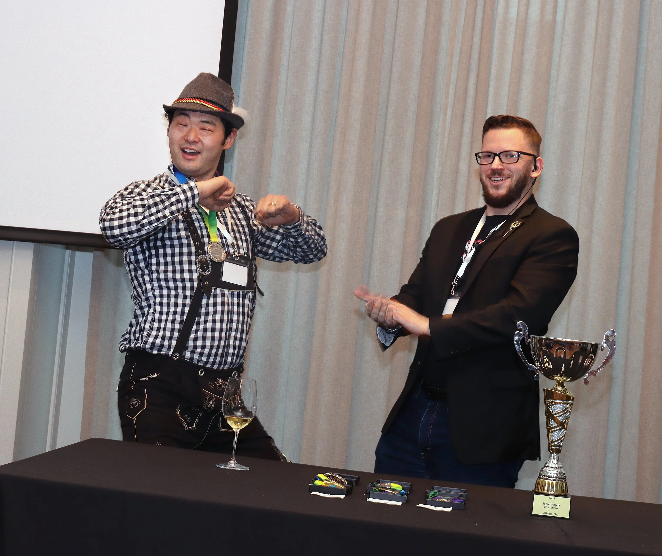 Two men celebrating at an award ceremony. The man on the left is wearing a checkered shirt, hat, and medals, and is flexing his muscles. The man on the right is wearing a suit and glasses, smiling, and has his arm around the first man. There is a lar