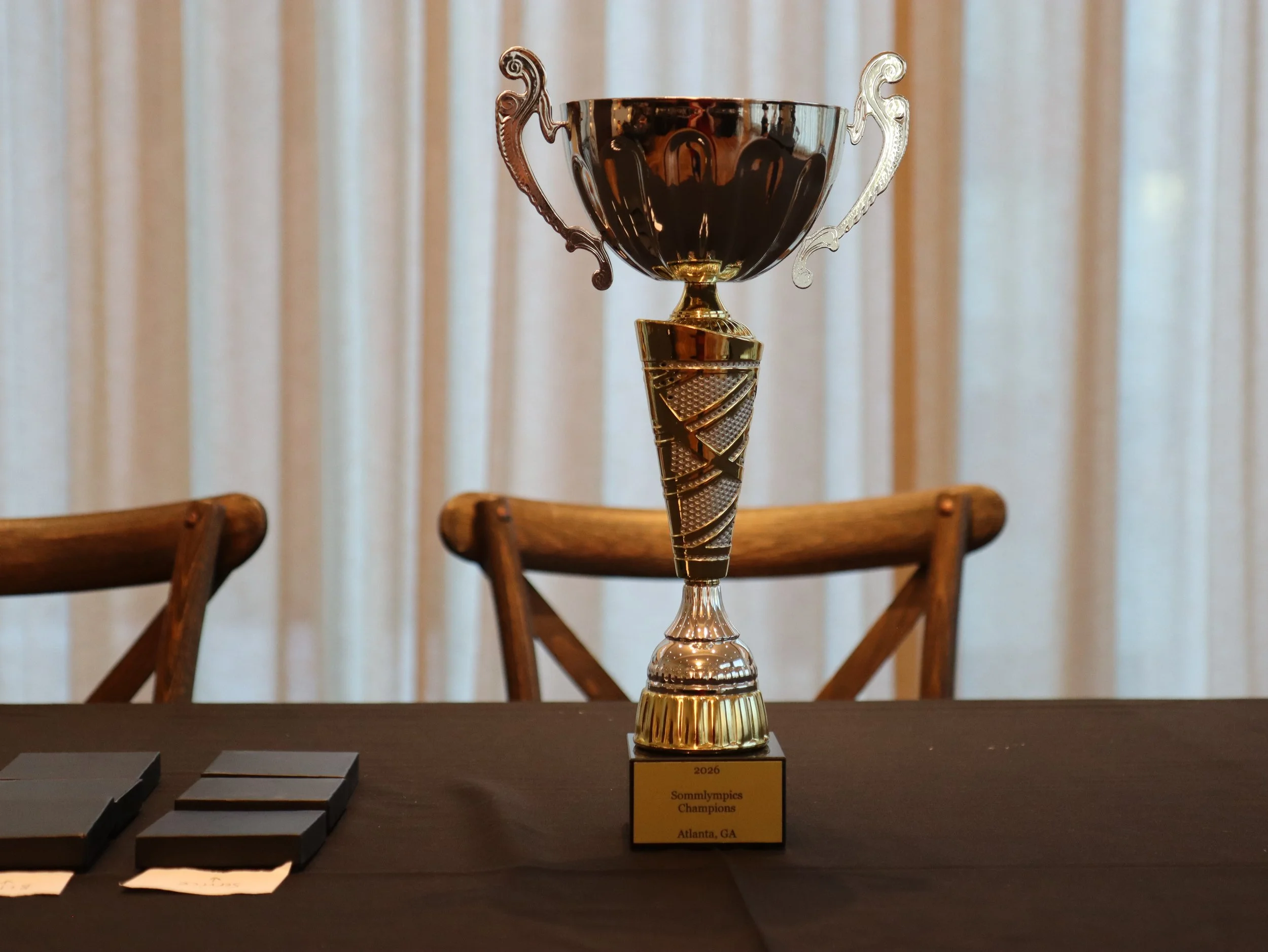 A trophy with a gold and silver design, placed on a table with a sign reading "2026 Schwimmypics Champions Atlanta, GA" in front of it. Part of a chessboard and chairs are visible in the background.