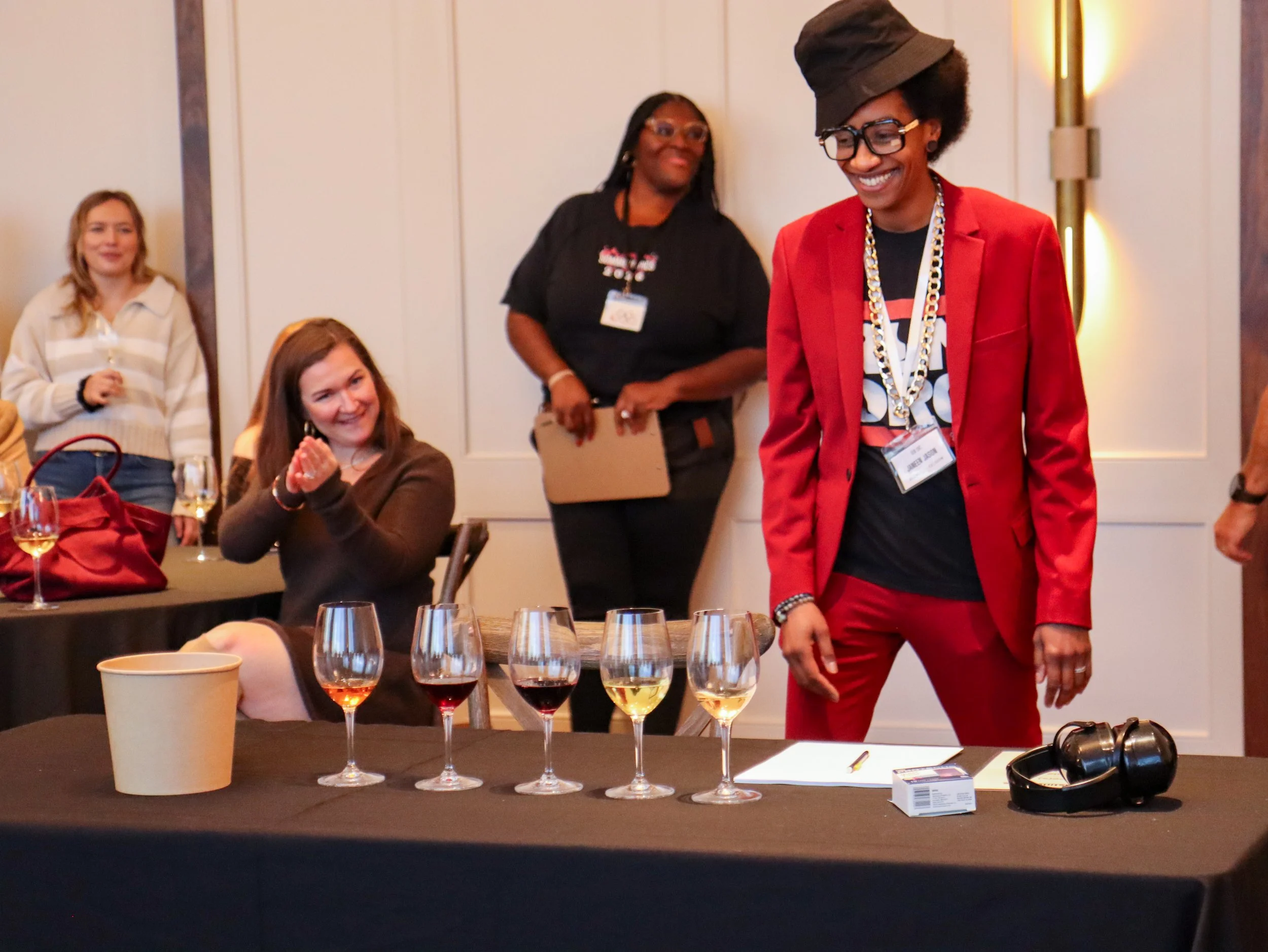 People at a table with wine glasses, a paper cup, and a headphone on a dark tablecloth, with some attendees standing in the background.