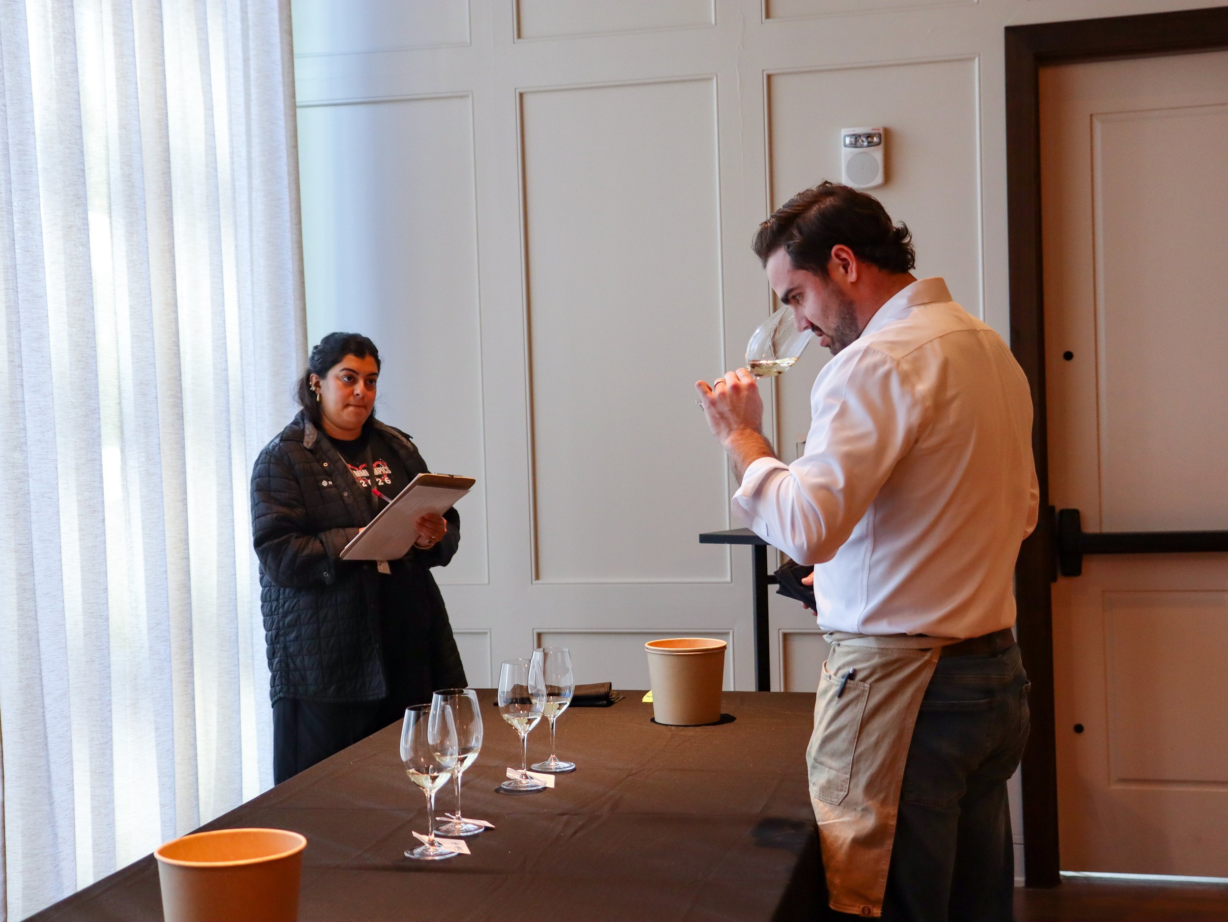 A man tasting white wine, a woman holding a clipboard, and wine glasses on a table in a room with beige walls and white curtains.