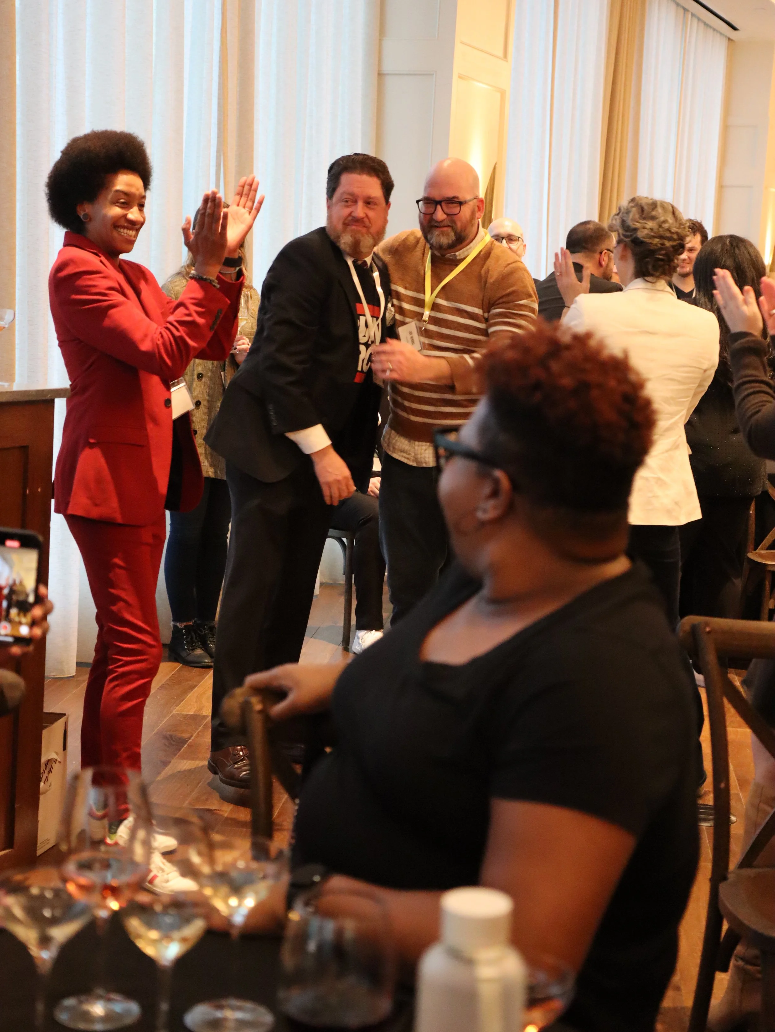 People at a social gathering or party, with some standing, smiling, and talking. A woman in a red suit is waving or greeting, and a man in a black jacket is interacting with another man in a striped sweater. The background shows other guests and curt