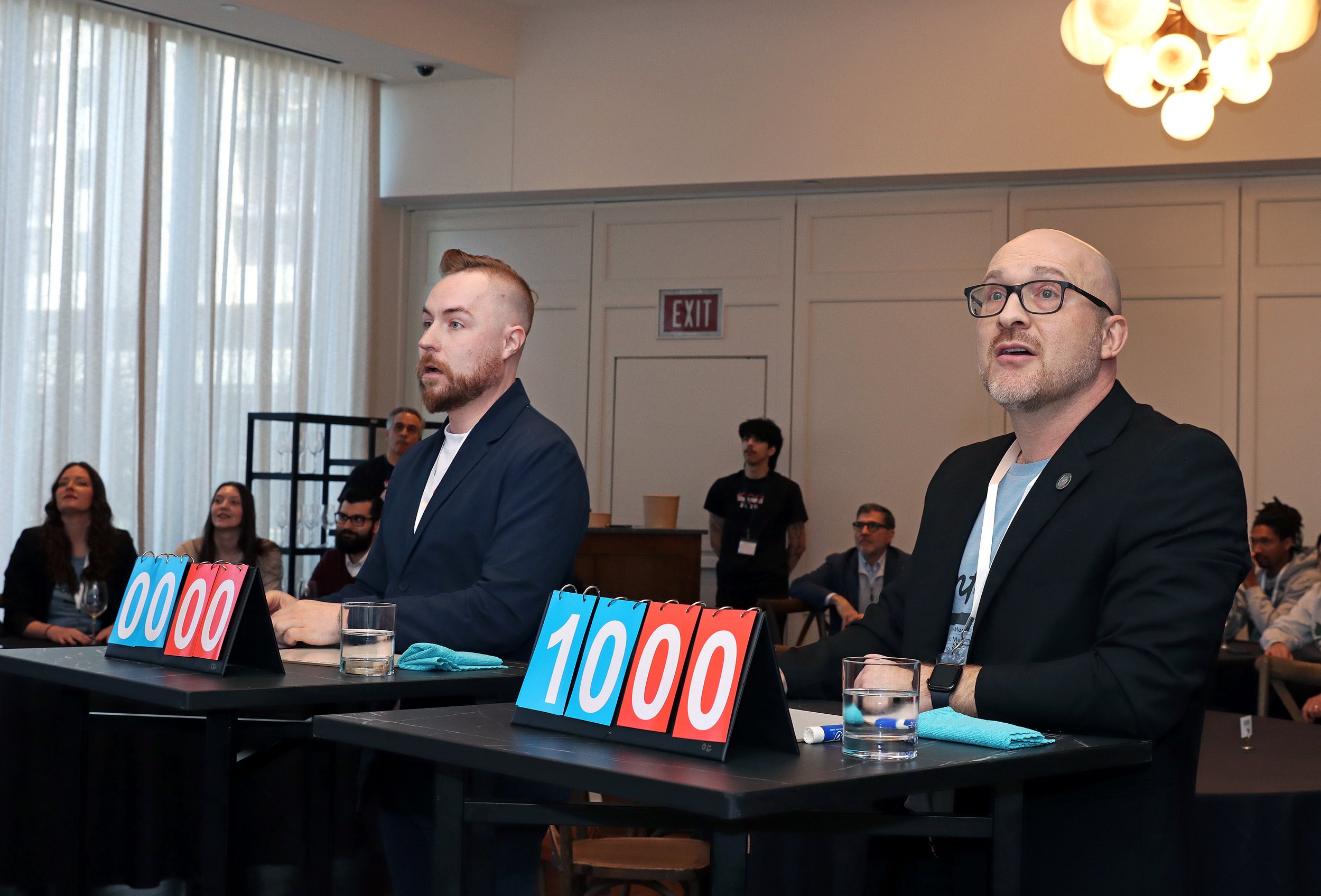 Two men sitting at a table during a game show or competition, with scoreboards displaying 000, 100, and 200 points, in a well-lit conference room with a chandelier and an exit sign in the background. Several audience members are visible in the backgr