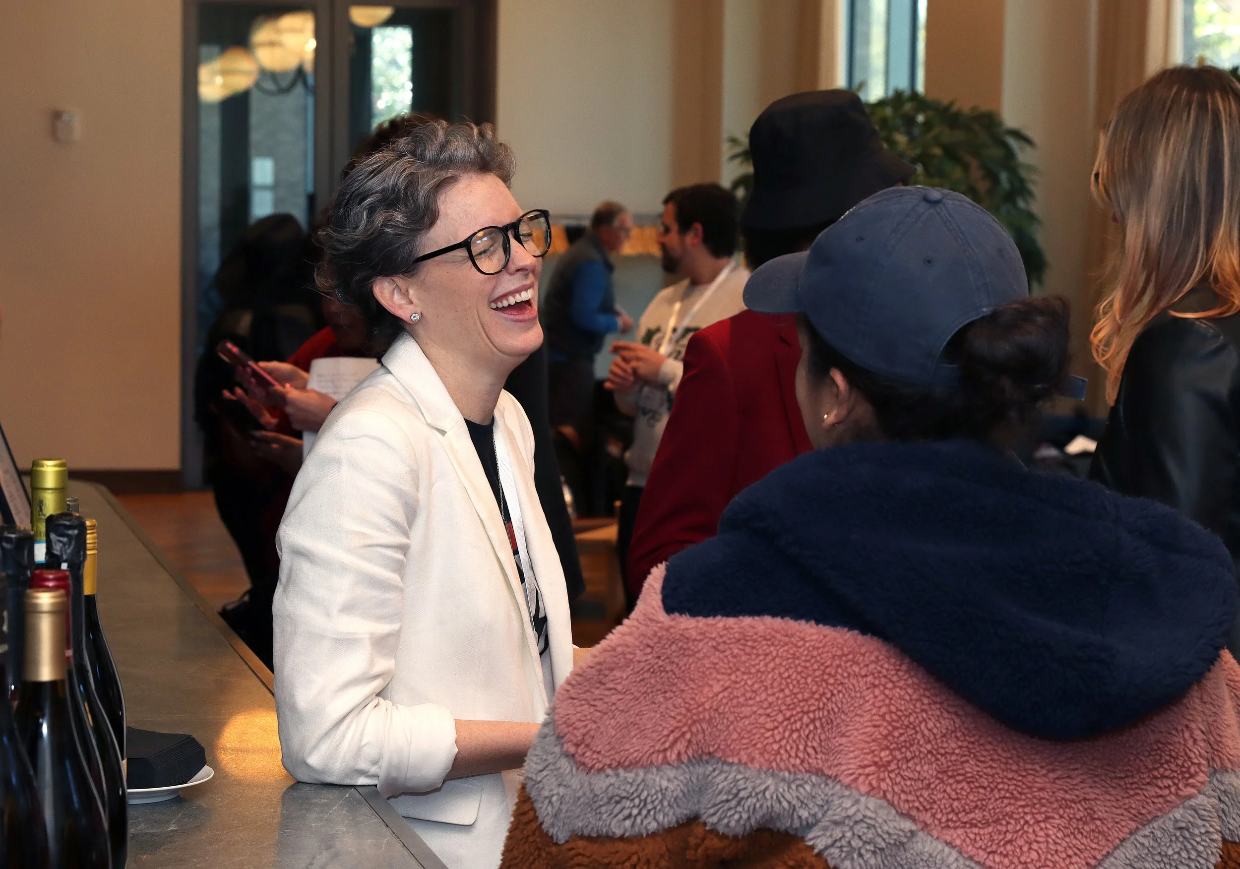 A group of people socializing inside a modern building with large windows, including a woman with short gray hair, glasses, wearing a white blazer, smiling and talking to others near a bar with bottles in the foreground.
