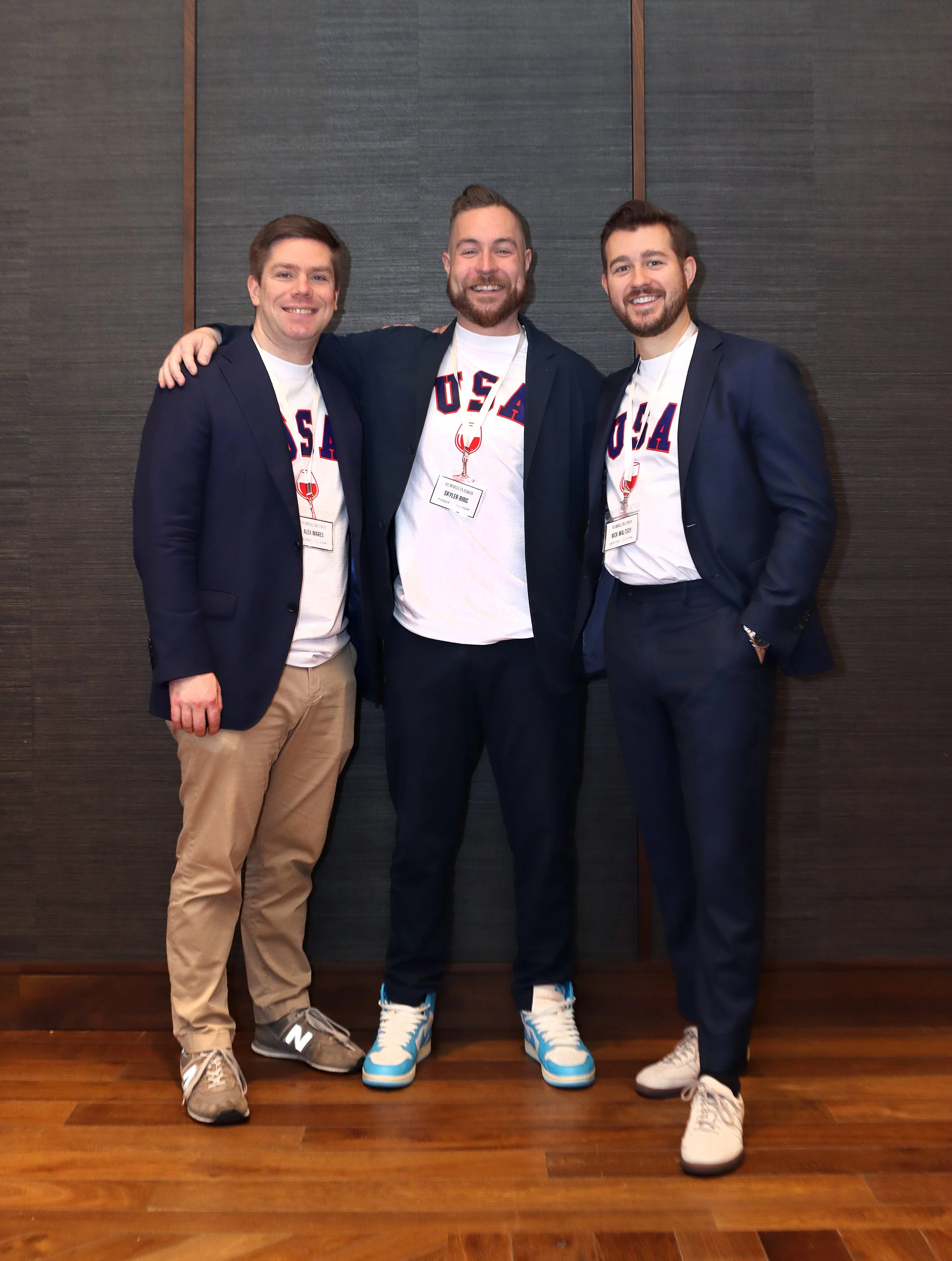 Three young men in suits with USA-themed t-shirts underneath, standing closely together and smiling for the camera in front of a dark textured wall. All are wearing casual sneakers and have name tags around their necks.