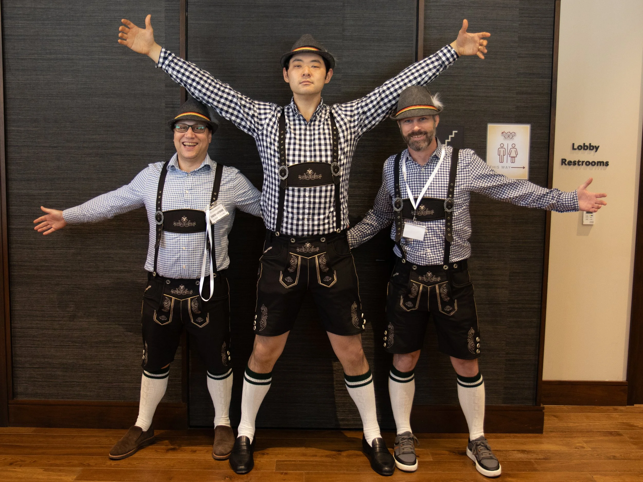 Three men dressed in traditional Bavarian-style clothing, wearing checkered shirts, lederhosen, and hats, standing in front of a dark wall with their arms raised in excitement.