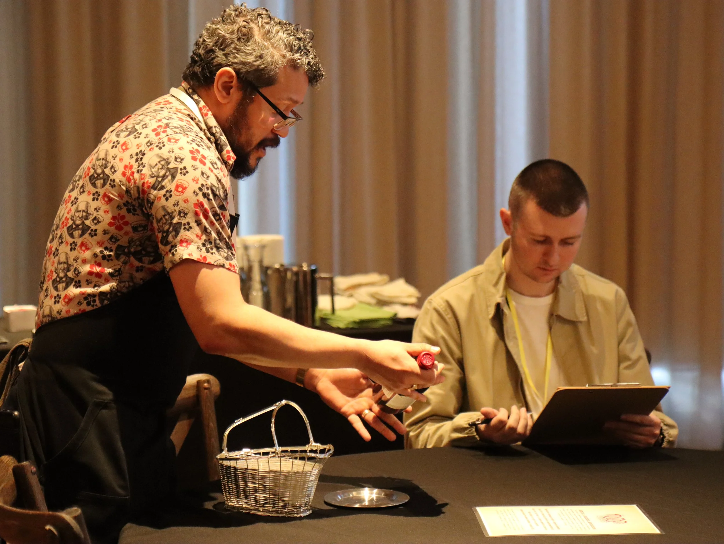 A man with curly hair and glasses in a patterned shirt is leaning over a seated young man with short hair, wearing a beige jacket, in a room with brown curtains. They appear to be engaged in some sort of activity or conversation, with a picnic basket