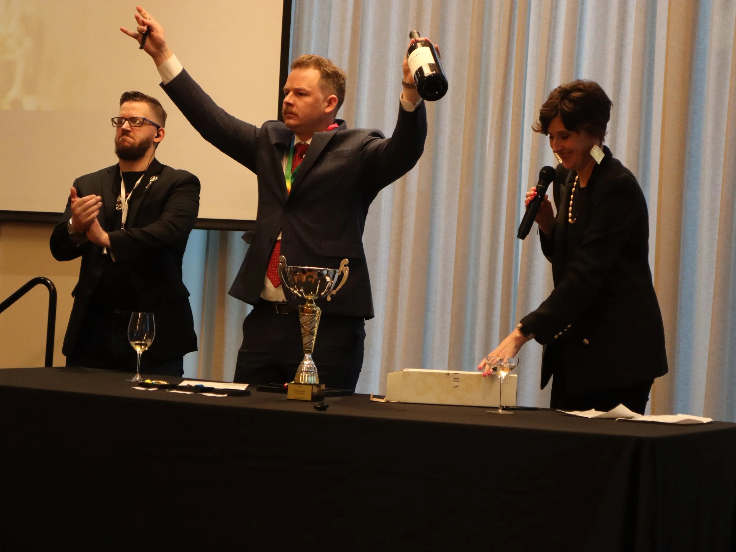 Three people dressed in formal attire are on a stage, with a table in front of them holding a large trophy, a wine glass, and a box. One man, in the center, is raising a wine bottle in one hand and a pen in the other. The woman on the right is smiling and holding a microphone and a glass of wine. The man on the left appears to be clapping or about to clap.