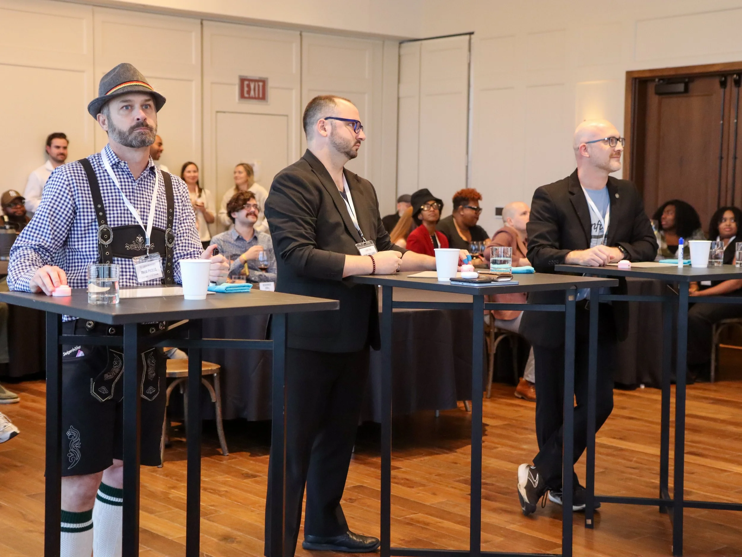 Three men sitting at a table during a conference or event, attentively listening, with additional attendees visible in the background.