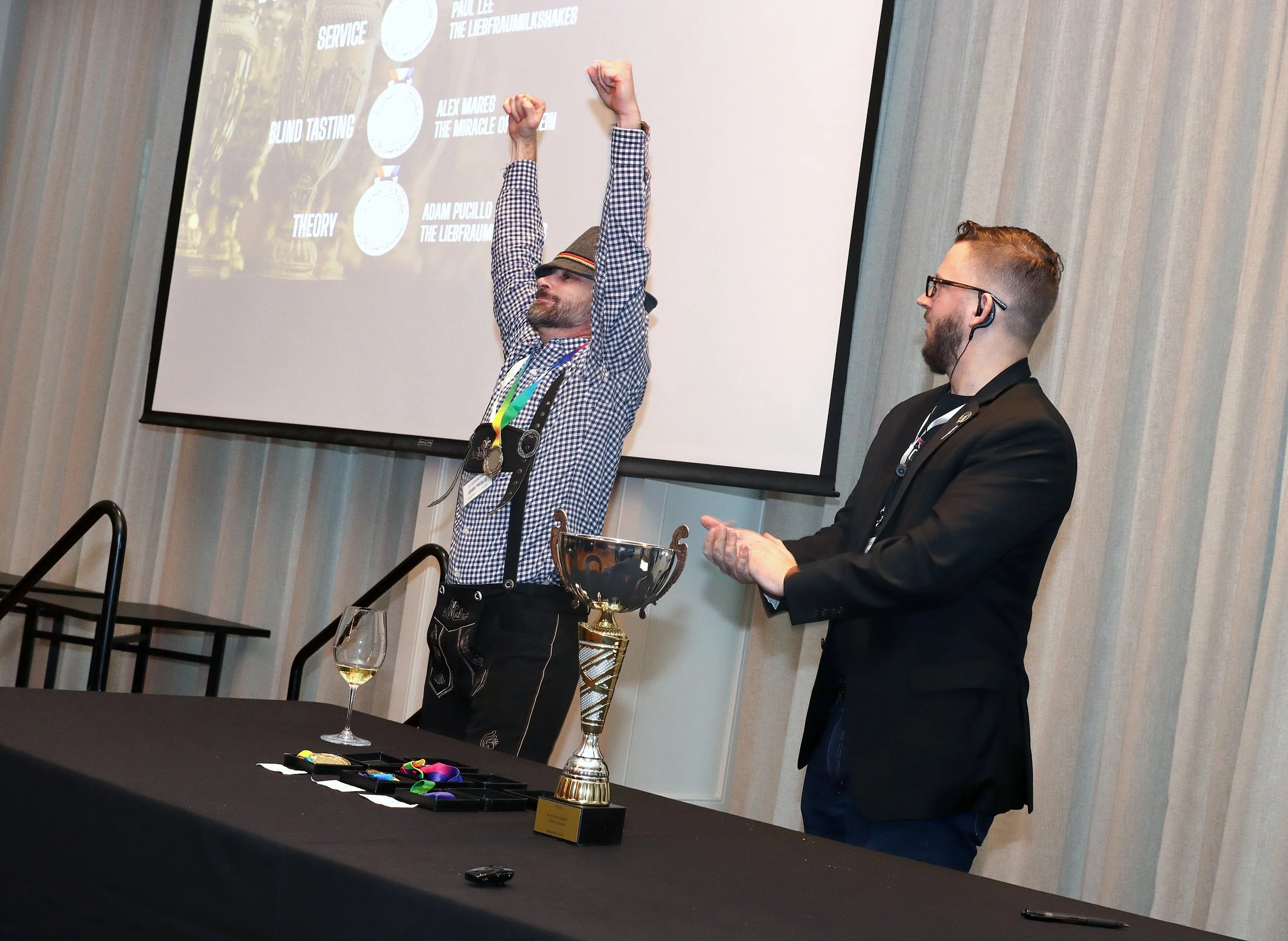 Two men at an award ceremony or competition standing behind a table with trophies, medals, and ribbons, one holding a large silver trophy. A large screen in the background displays text and images related to a science or academic event.
