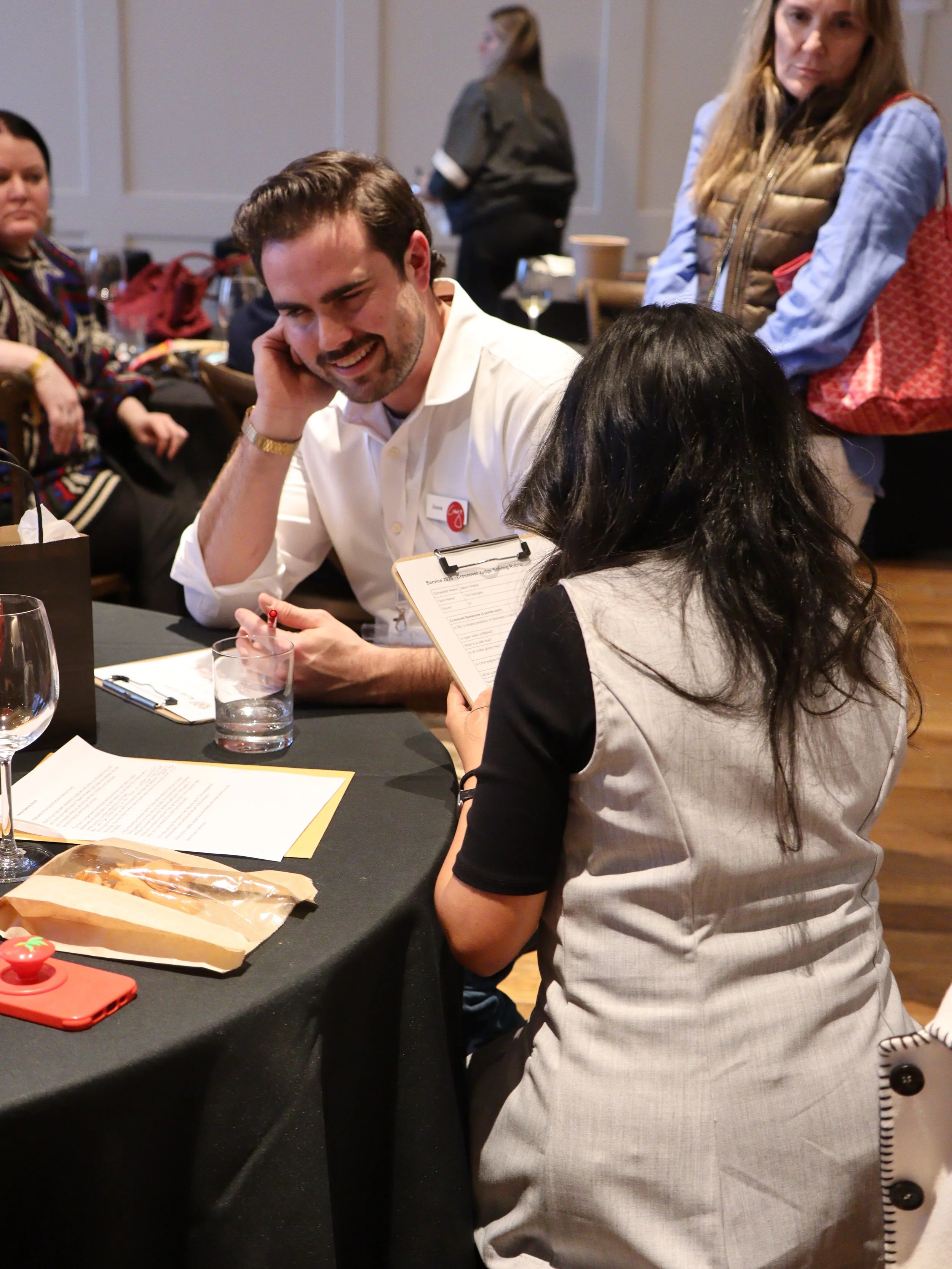 A group of people sitting and standing at a social gathering or event, with some engaging in conversation and smiling. The setting appears to be indoors with a table, documents, and snacks visible.