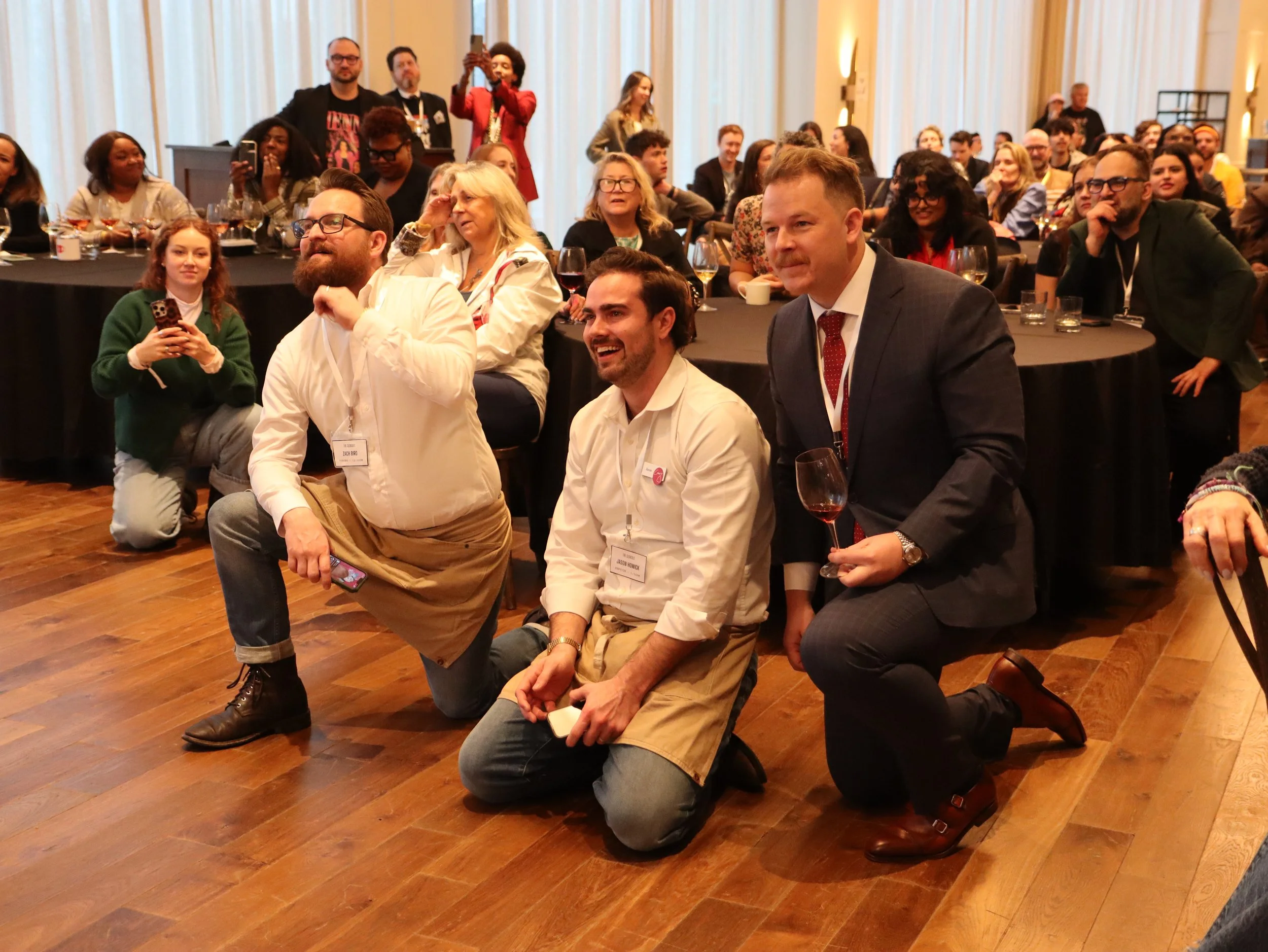 A diverse group of people at a conference or event, sitting and kneeling on the floor, with some holding drinks and others taking pictures or looking at the stage. The background shows large curtains and a speaker at a podium.