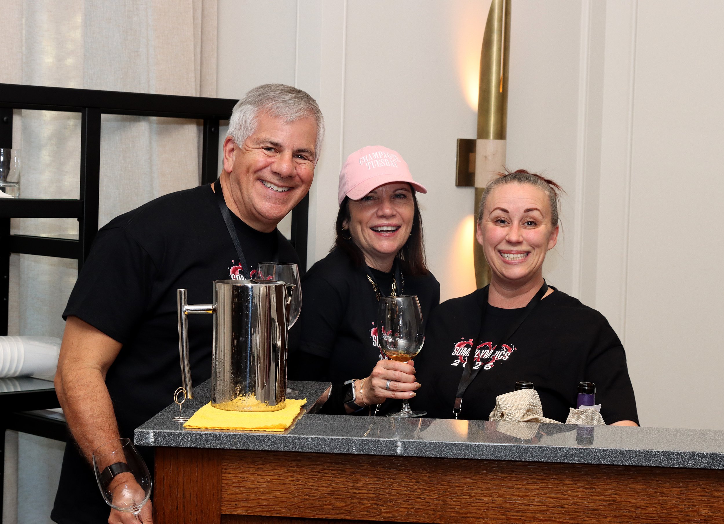 Three smiling people standing behind a bar, with glasses of wine in their hands, celebrating at an indoor event. They are dressed casually, and there are wine bottles and a champagne bucket on the bar in front of them.