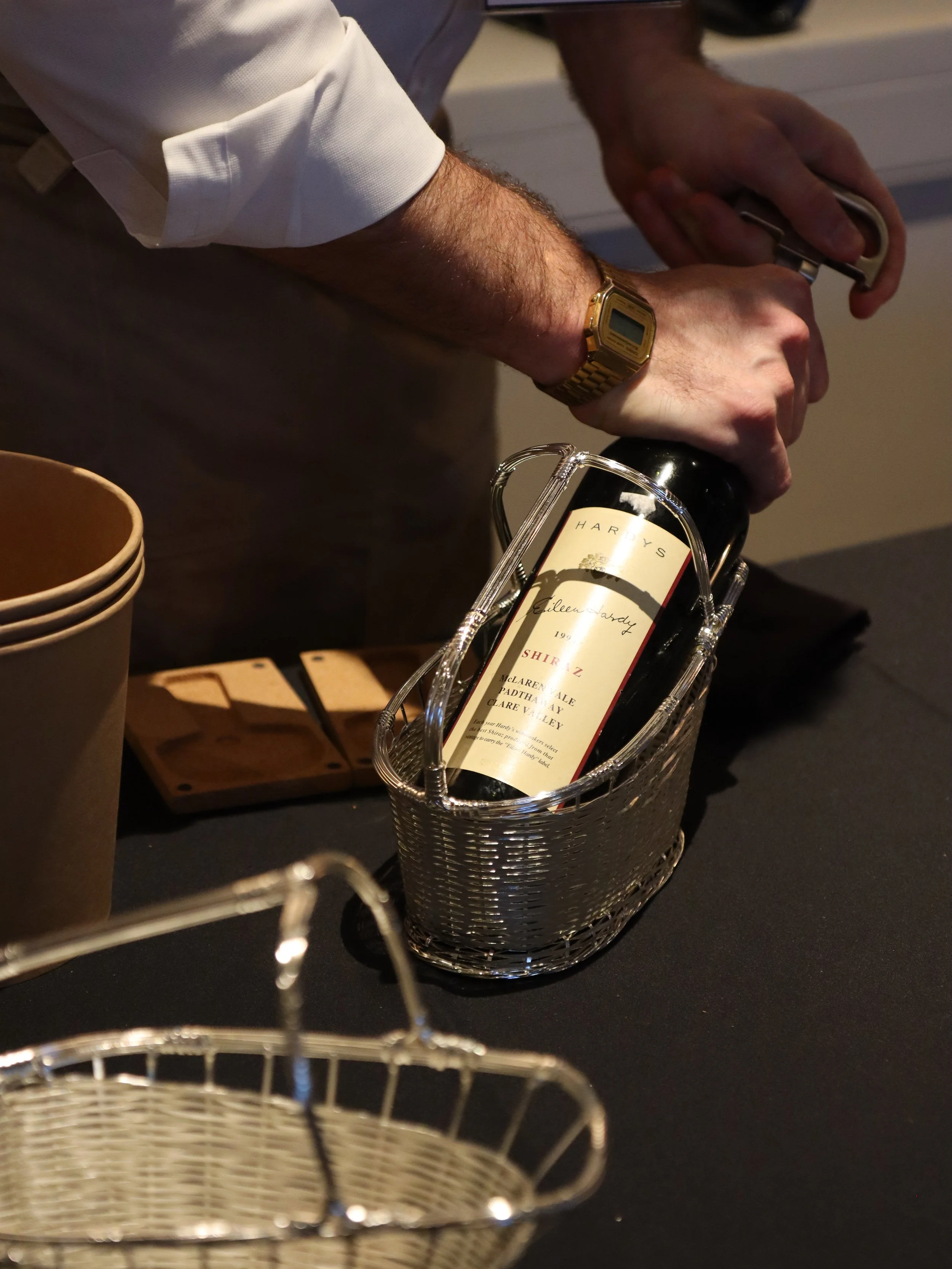 A person with hairy arms, wearing a white long-sleeve shirt and a gold digital watch, is opening a bottle of wine in a silver wire basket. There are beige bowls and a wooden tray nearby on a black surface.