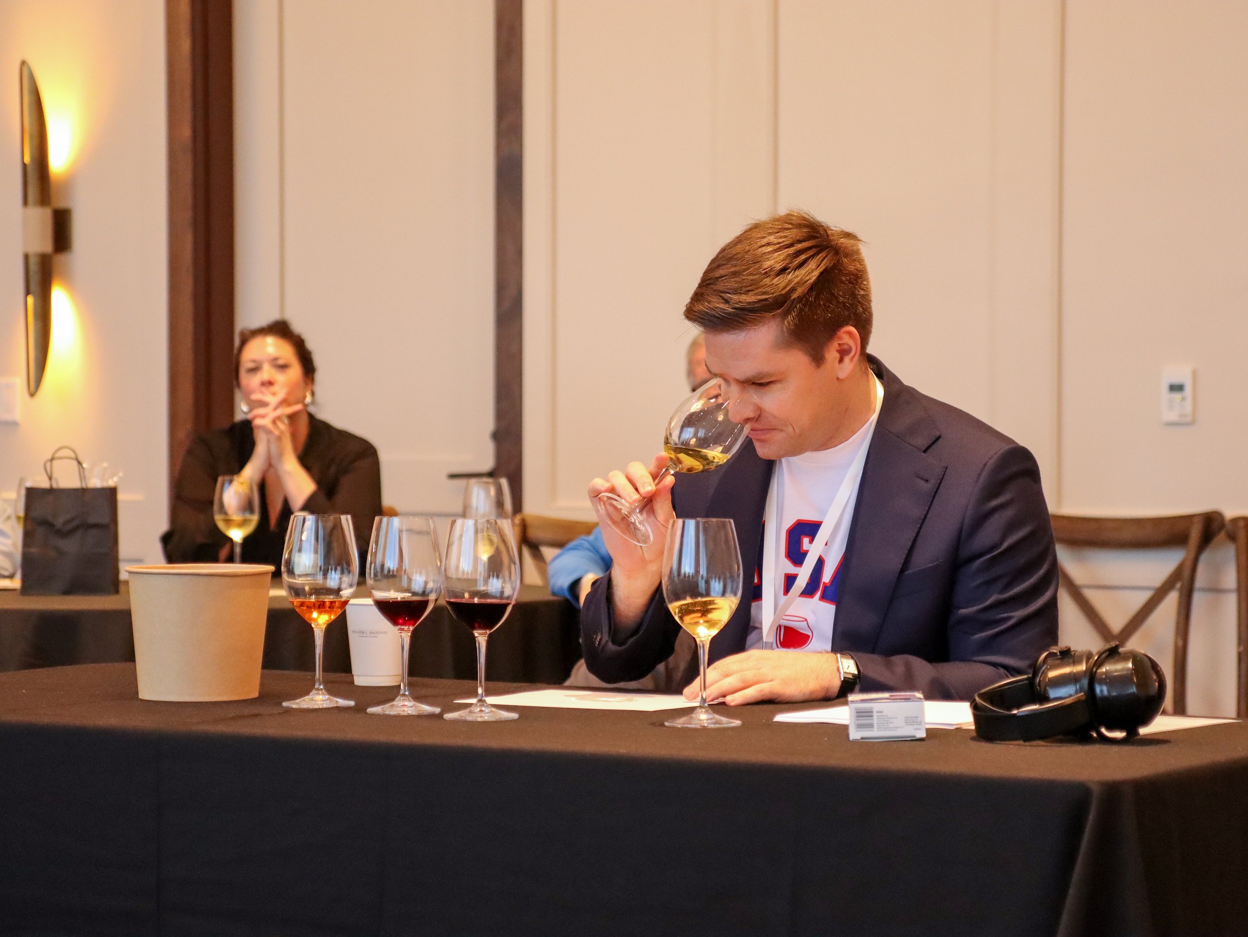 A man in a suit and a woman in a black outfit sitting at a table with wine glasses, tasting white wine, in a conference or meeting room with beige walls and wood paneling.