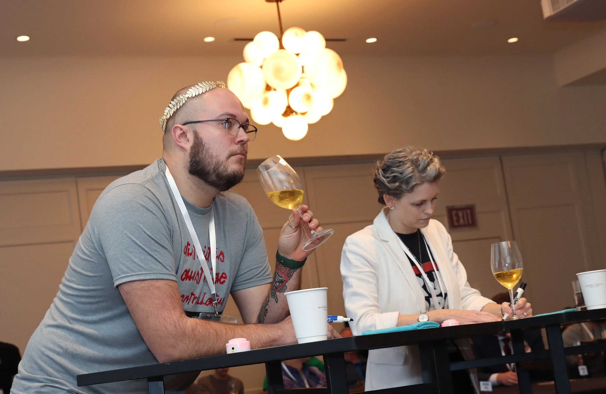 A man with glasses and a beard, wearing a grey T-shirt with red lettering, is sitting at a table holding a glass of white wine. Next to him, a woman with short curly hair, wearing a white blazer and black shirt, is also sitting at the table. They are