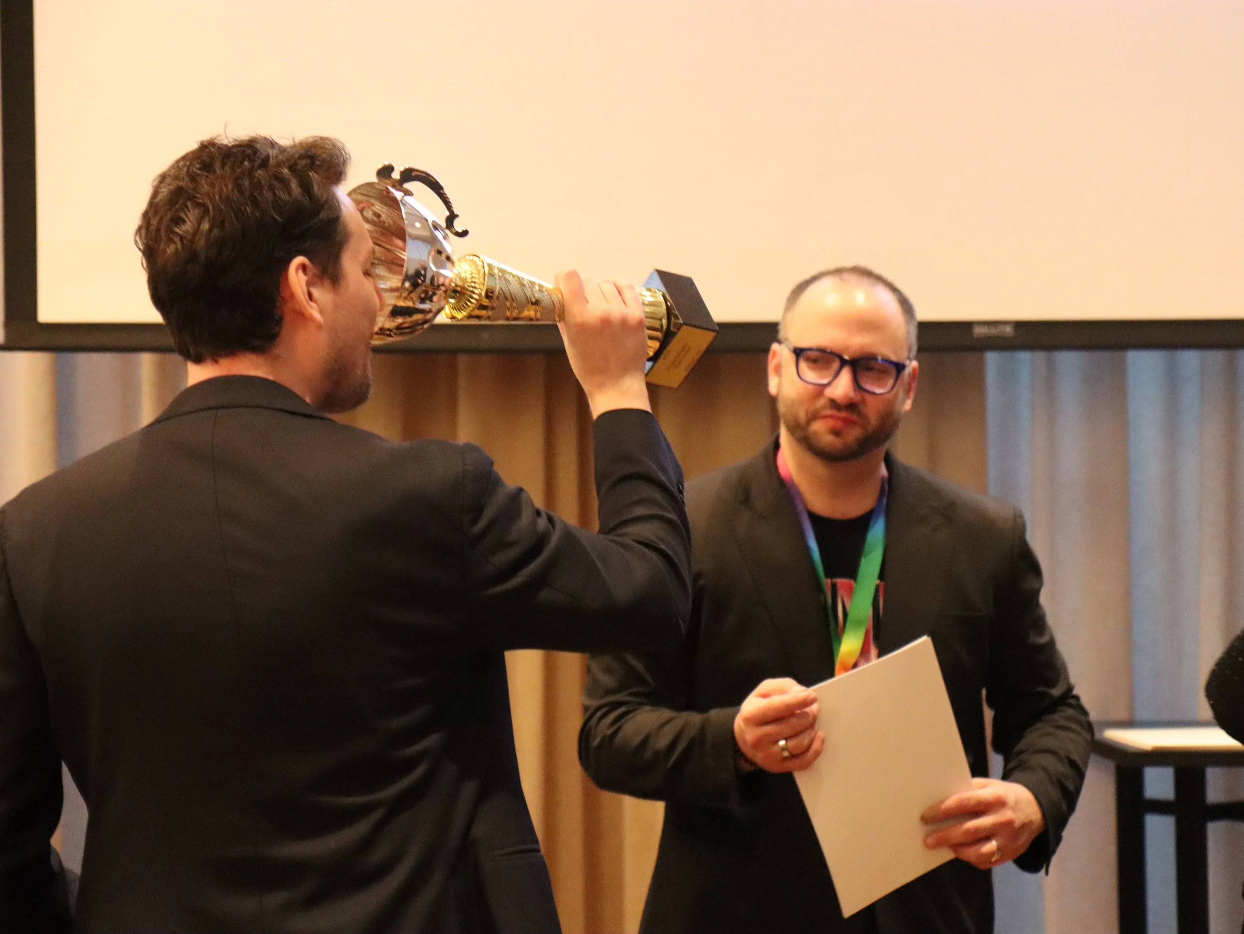 A man in a black suit and another man with glasses and a black blazer are at an award ceremony. The man in the suit is holding a trophy and appears to be presenting or about to present it to the man with glasses, who is holding a document or certific