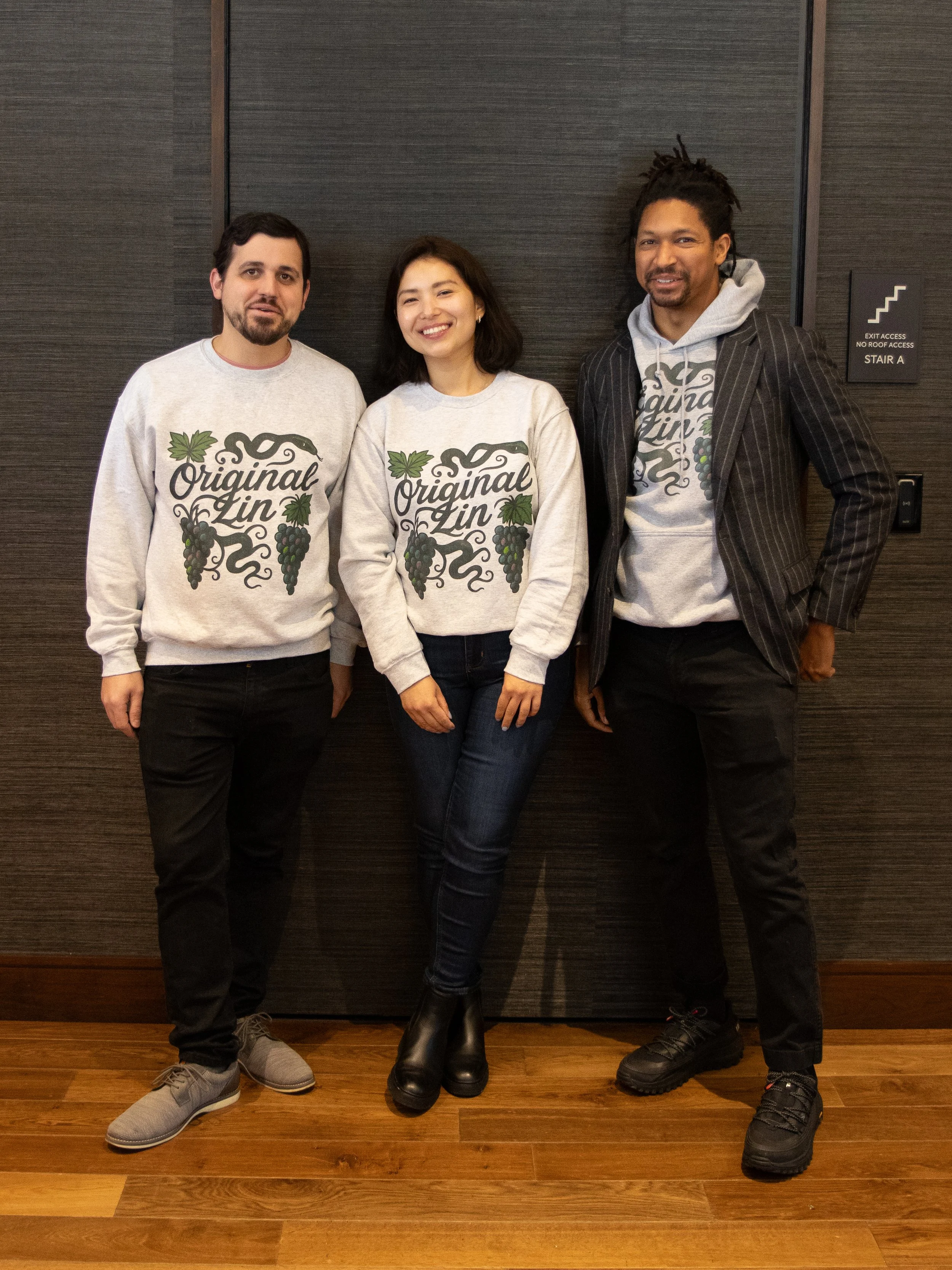 Three people standing in front of a dark wall, smiling and wearing sweatshirts that say 'Original Zin' with grapevine and grape cluster graphics. The person on the right is wearing a pinstripe blazer over a sweatshirt, and the woman is in the middle 