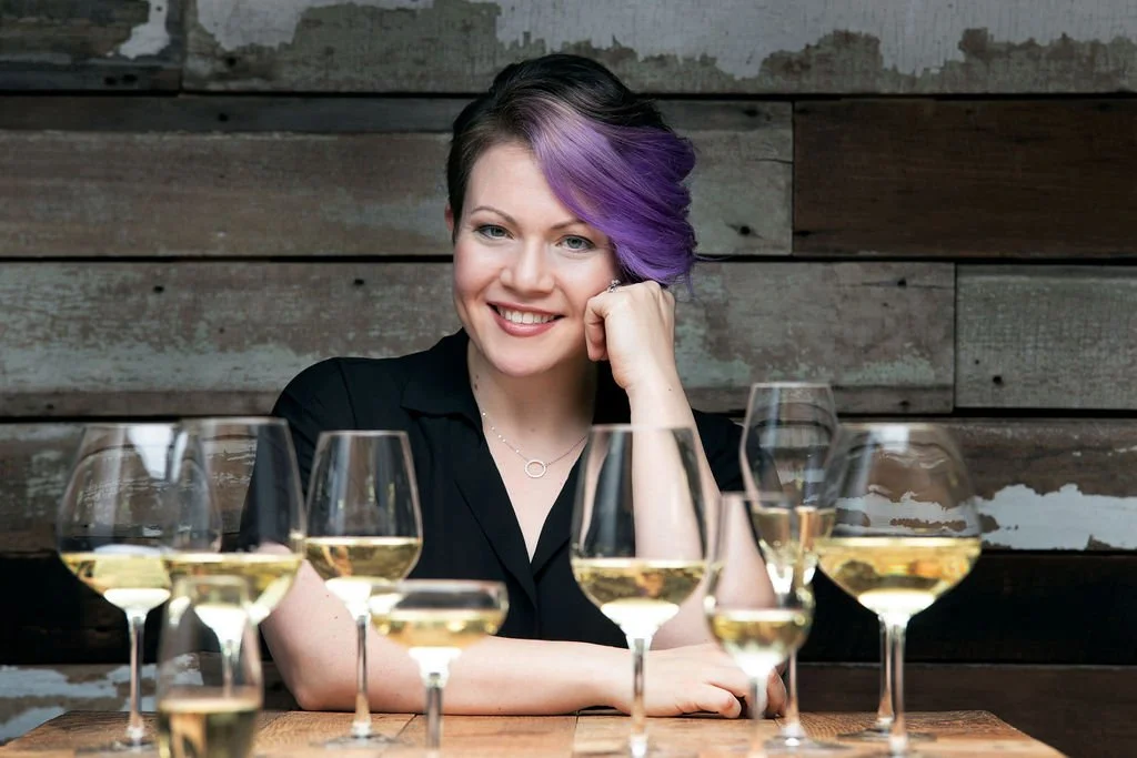 A woman with short purple hair is smiling at the camera while sitting at a table with several glasses of white wine.