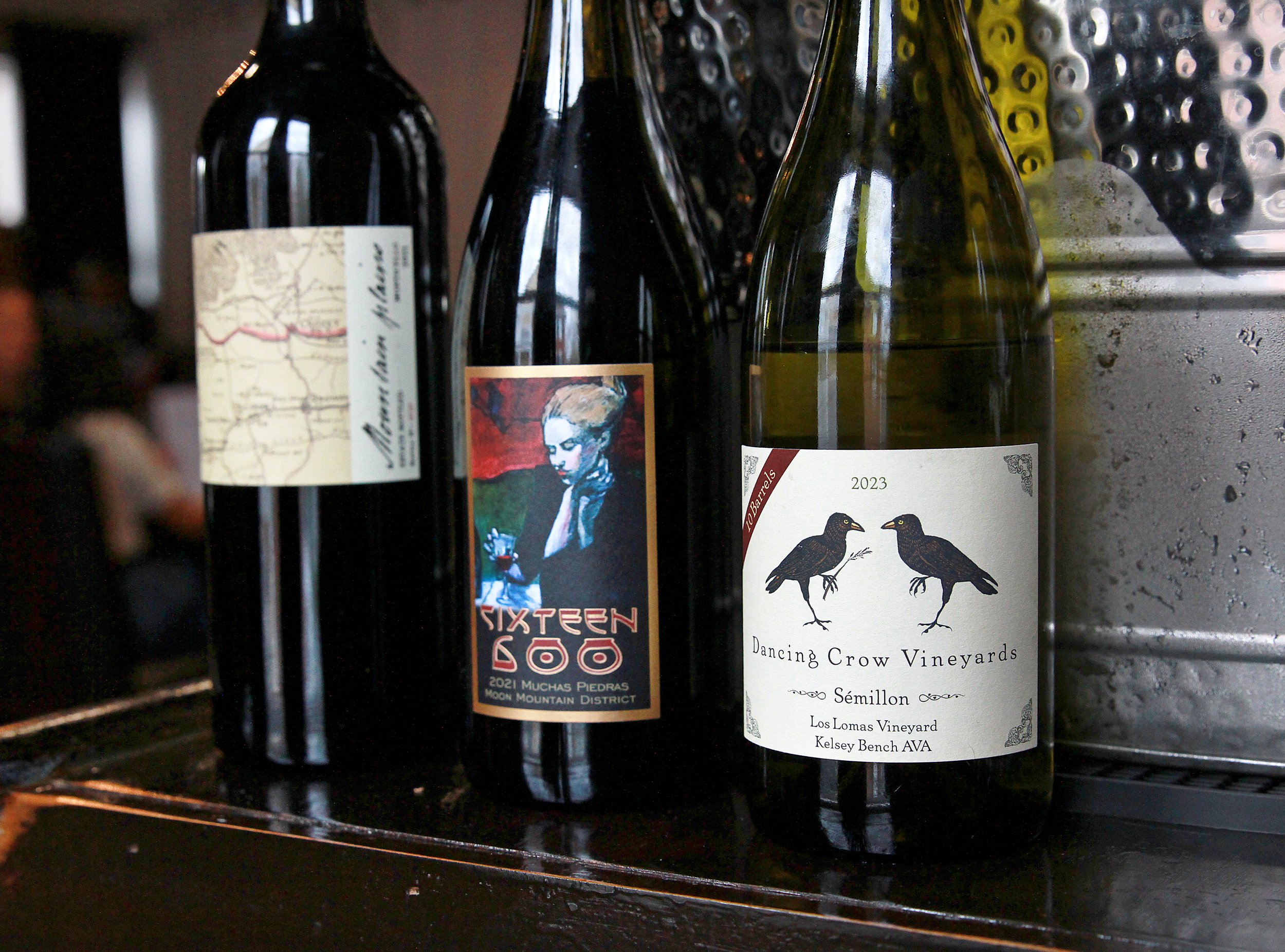 Three wine bottles on a dark surface in front of a metallic container with water droplets.