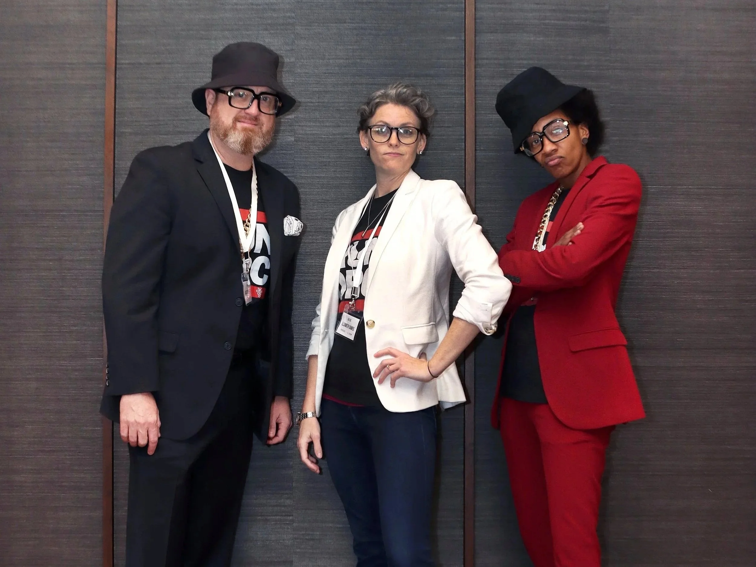 Three people standing in front of a dark wall. The person on the left is a man with a beard, glasses, a black hat, and a black blazer. The person in the middle is a woman with short hair, glasses, wearing a white blazer and a black shirt. The person on the right is a woman with short hair, glasses, a black hat, and a red blazer with her arms crossed.