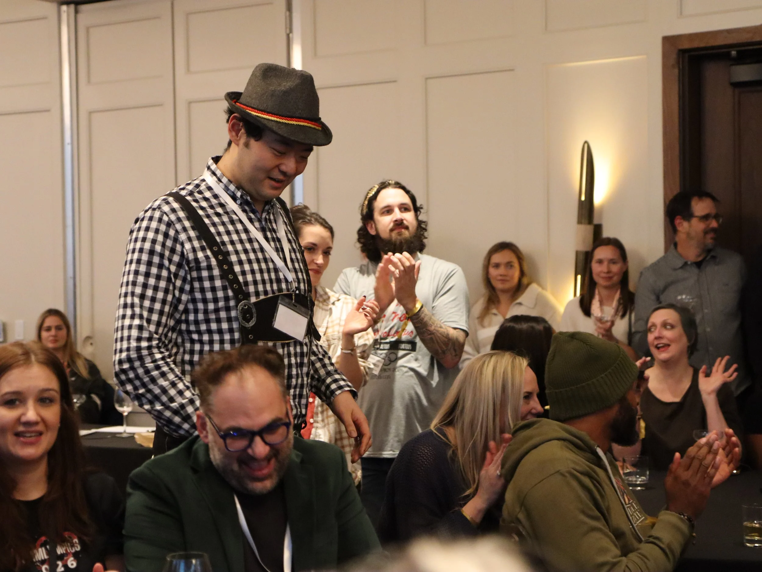 A group of people standing and sitting at a social gathering in a room with beige walls and modern wall lights. A man in a checkered shirt, gray hat, and camera around his neck is looking down. Others are clapping, smiling, and engaging in conversati