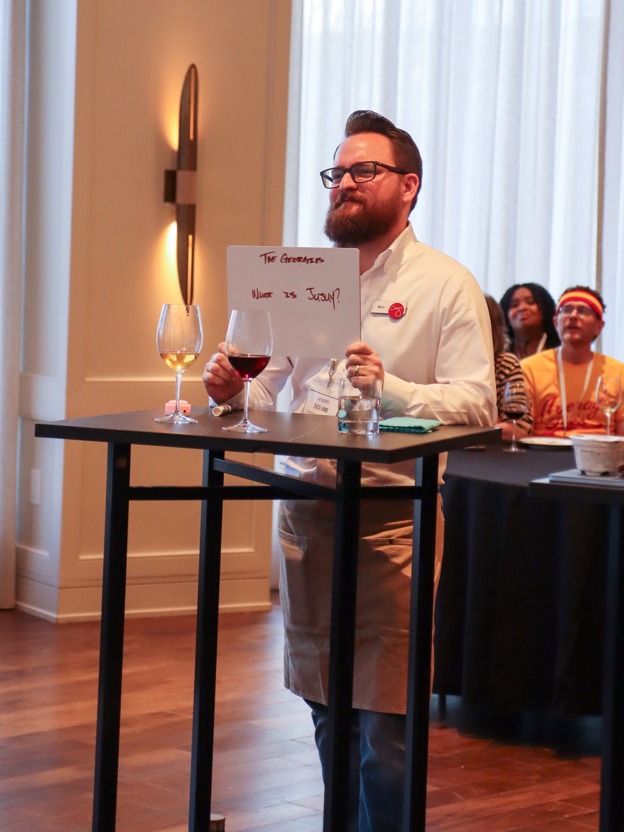 Man with glasses, beard, wearing a white shirt, standing behind a high table with two glasses of wine, holding a whiteboard with handwritten notes. There are people seated in the background at an indoor event.