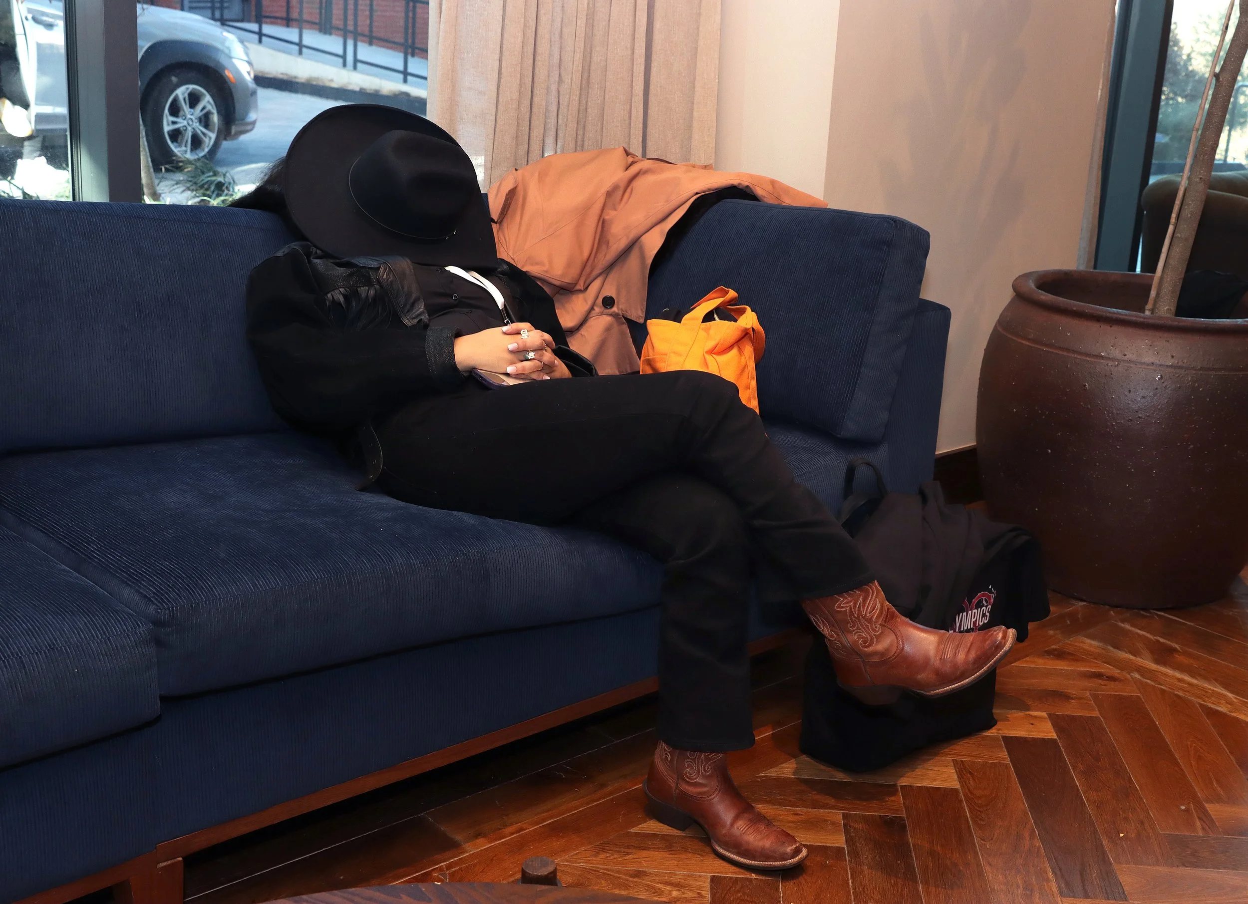 A woman with a black hat and black outfit is asleep on a blue couch in a room with wooden flooring. She has rings on her fingers and is holding a smartphone. There are clothing items, an orange bag, and cowboy boots beside her. The background shows w