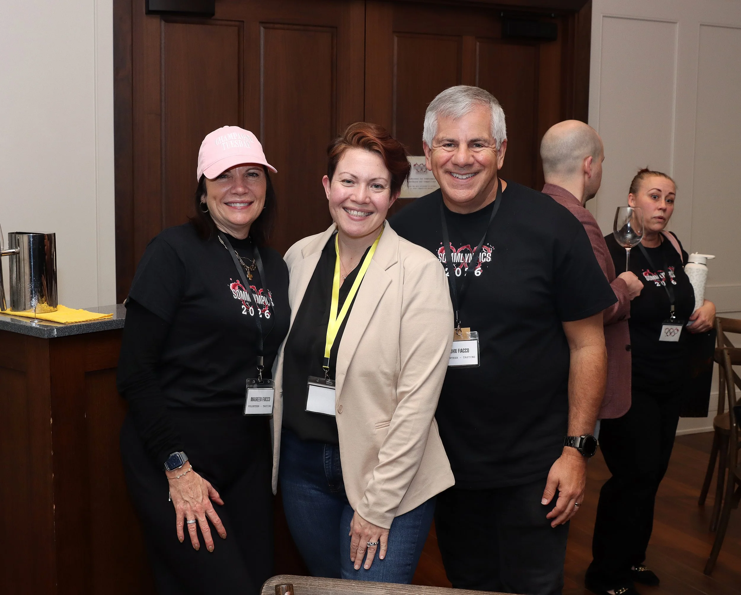 Group of people smiling at a social event indoors. Three women and one man in the foreground, with two women and a man in the background. The woman on the left is wearing a pink cap and black shirt, the woman in the middle is wearing a cream blazer, 