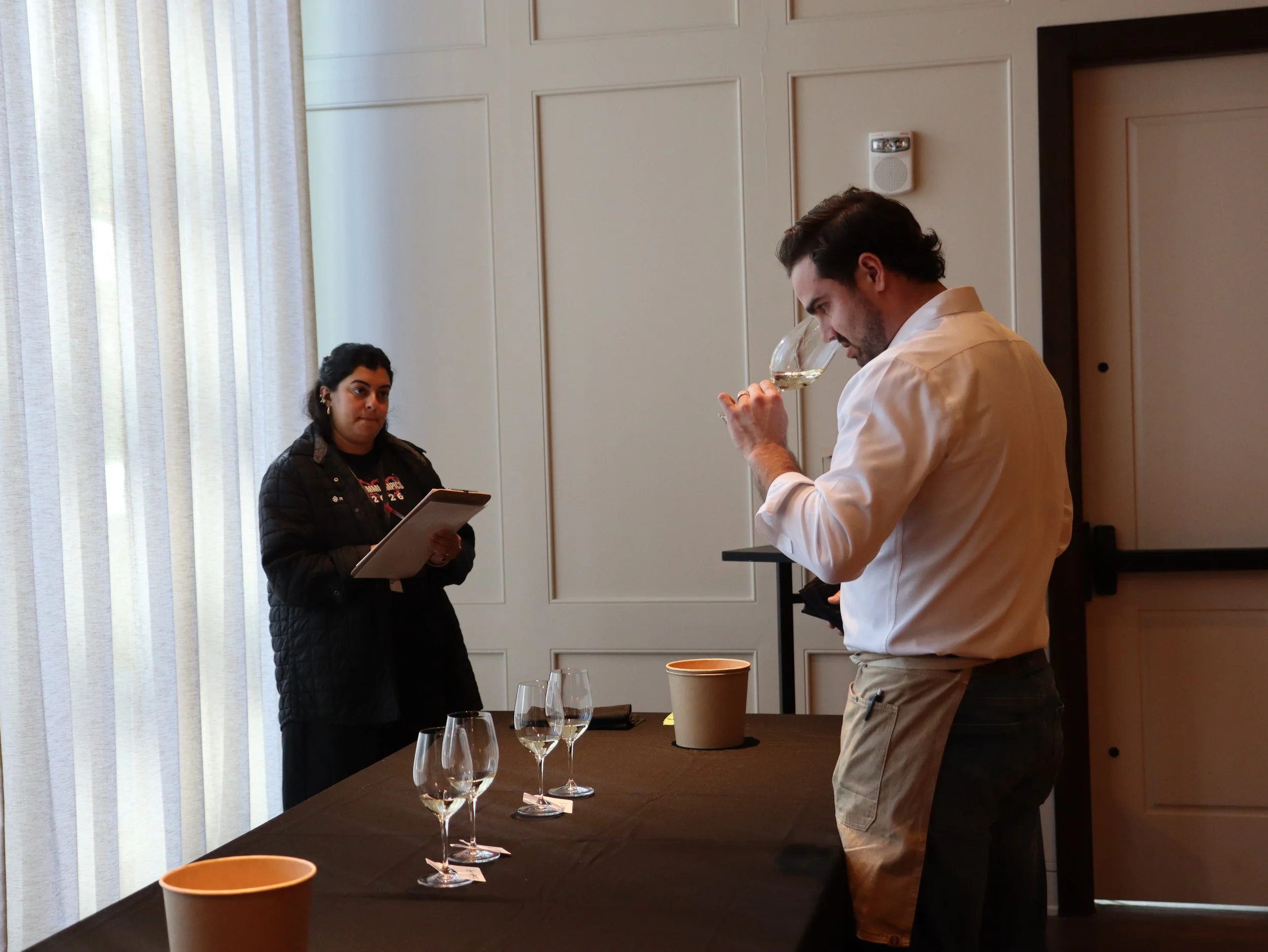 Two people are engaged in a wine tasting or evaluation. The man, in a white shirt and beige pants, smells a glass of white wine, while the woman, in a black jacket, holds a clipboard or notepad. Several wine glasses and disposable cups are on the tab