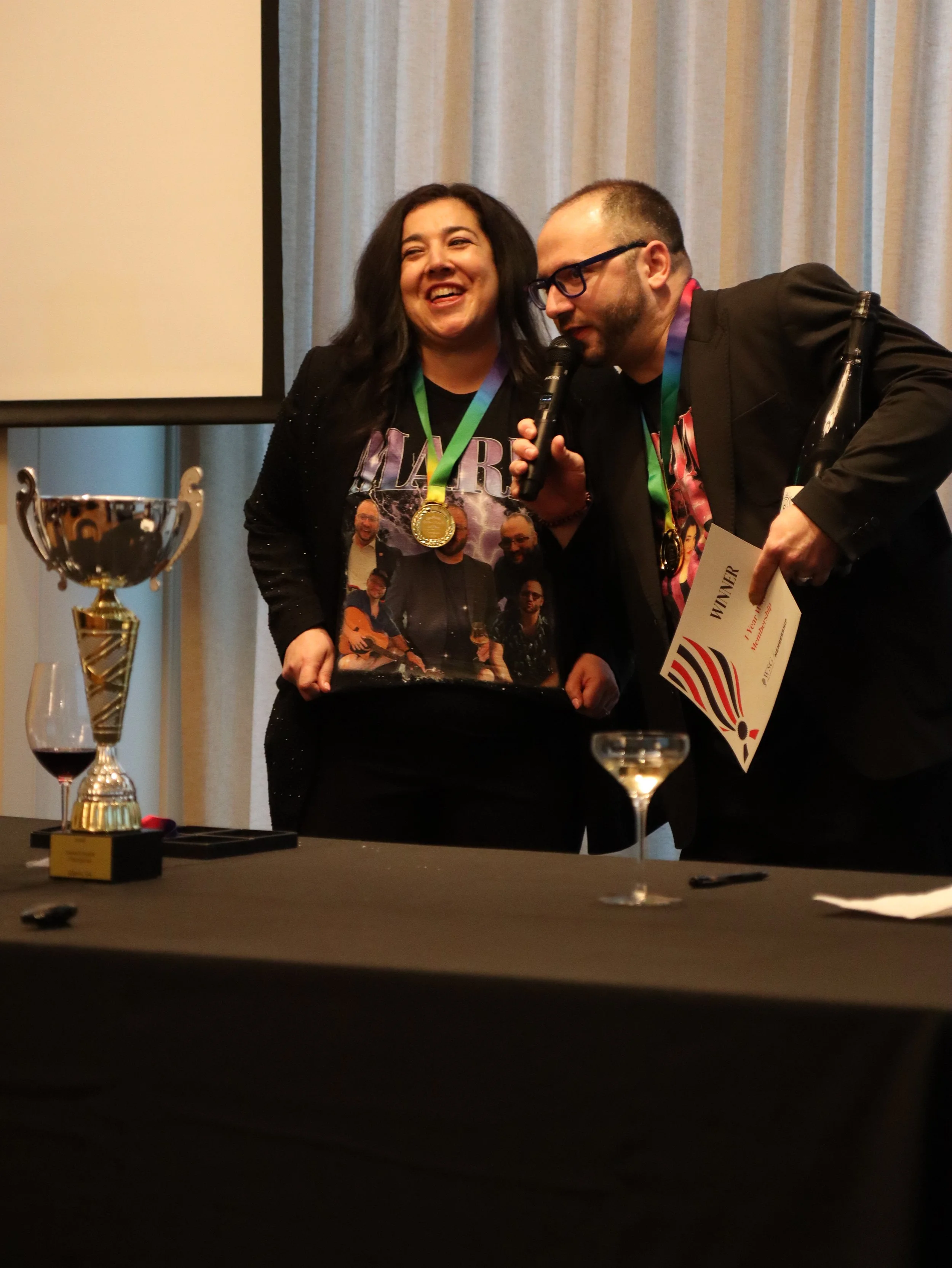 Two people, a woman wearing a black shirt with a band and a man with glasses, are smiling and speaking into a microphone at an awards ceremony. The woman is wearing a gold medal around her neck. There are trophies, a champagne glass, and a certificat