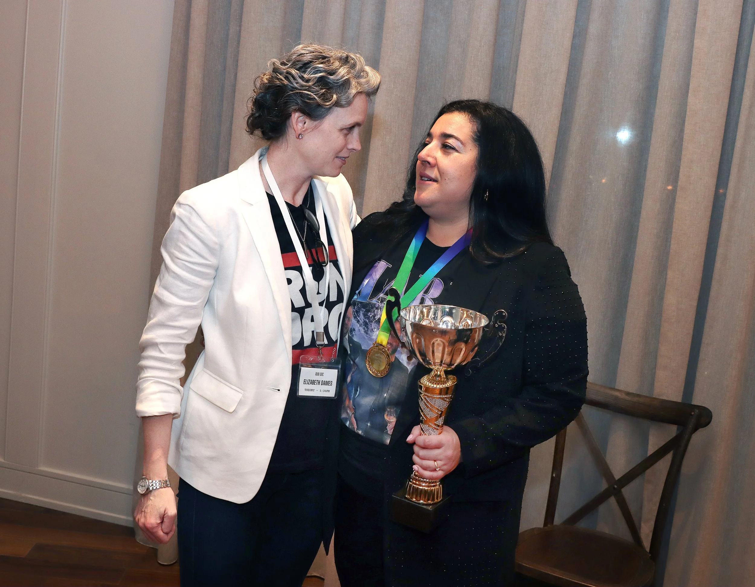 Two women standing close together, one holding a large gold trophy. One woman with short curly hair and wearing a white blazer is looking at the other woman with long black hair, who is wearing a black shirt with a colorful graphic and a purple ribbo