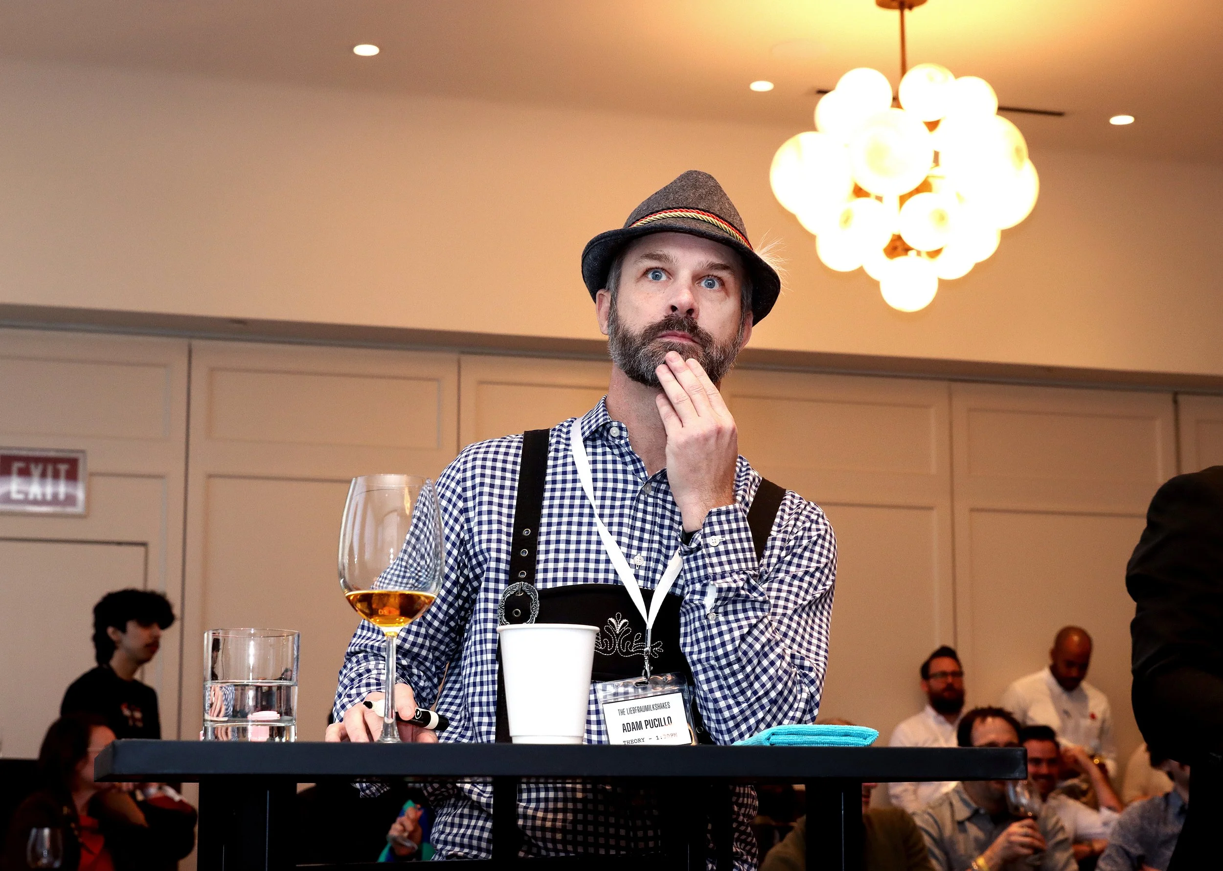 A man with blue eyes and a beard wearing a traditional German hat and checkered shirt, standing behind a table with wine glasses, a white cup, and a name tag, in a large room with other people in the background and a chandelier overhead.