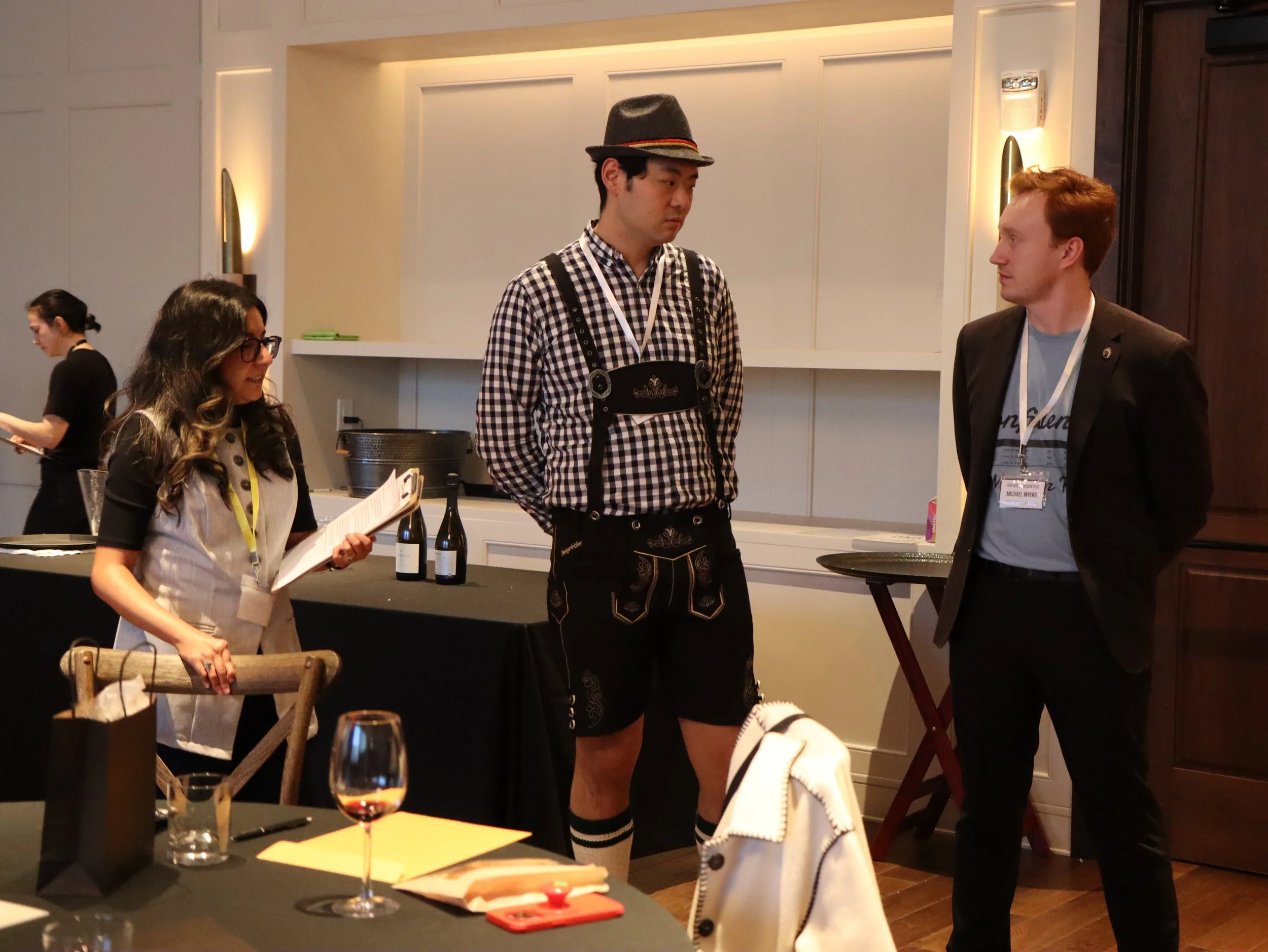 People socializing at a gathering with wine glasses and bottles on a table, a woman reading a menu, two men engaged in conversation, one in traditional German attire and the other in business casual, in a modern interior setting.