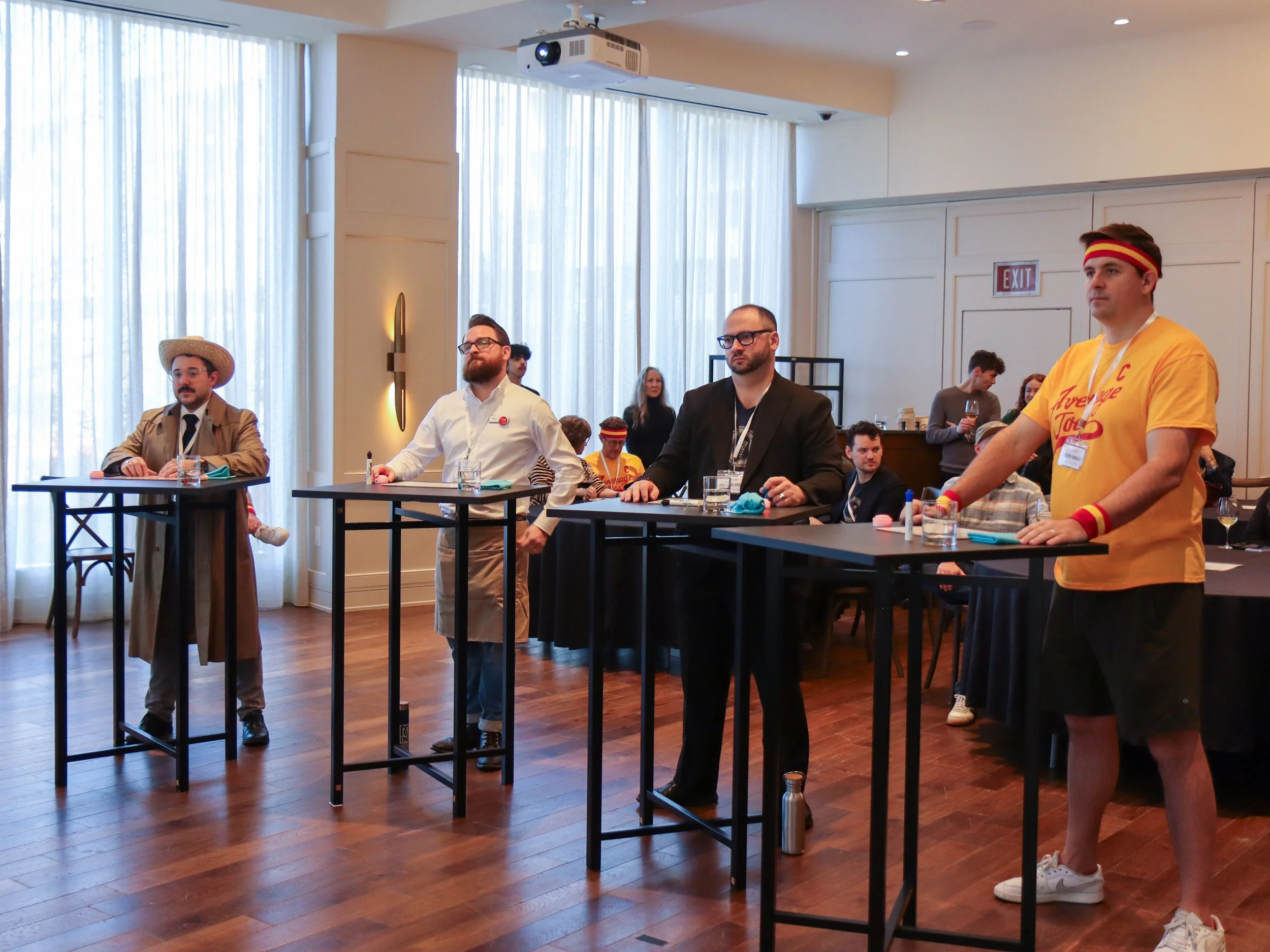 A group of people attending a conference or meeting in a well-lit room with tall curtains, some are sitting at tables while others are standing. Most of them are dressed in casual or business casual attire, with a few wearing themed or humorous acces