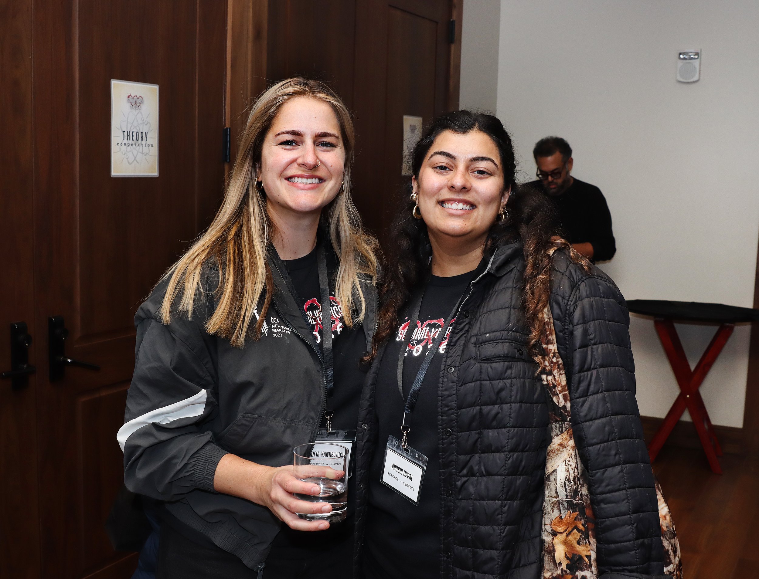 Two smiling women wearing conference name tags, one holding a glass of water, standing in a room with wooden paneling and a man in the background.