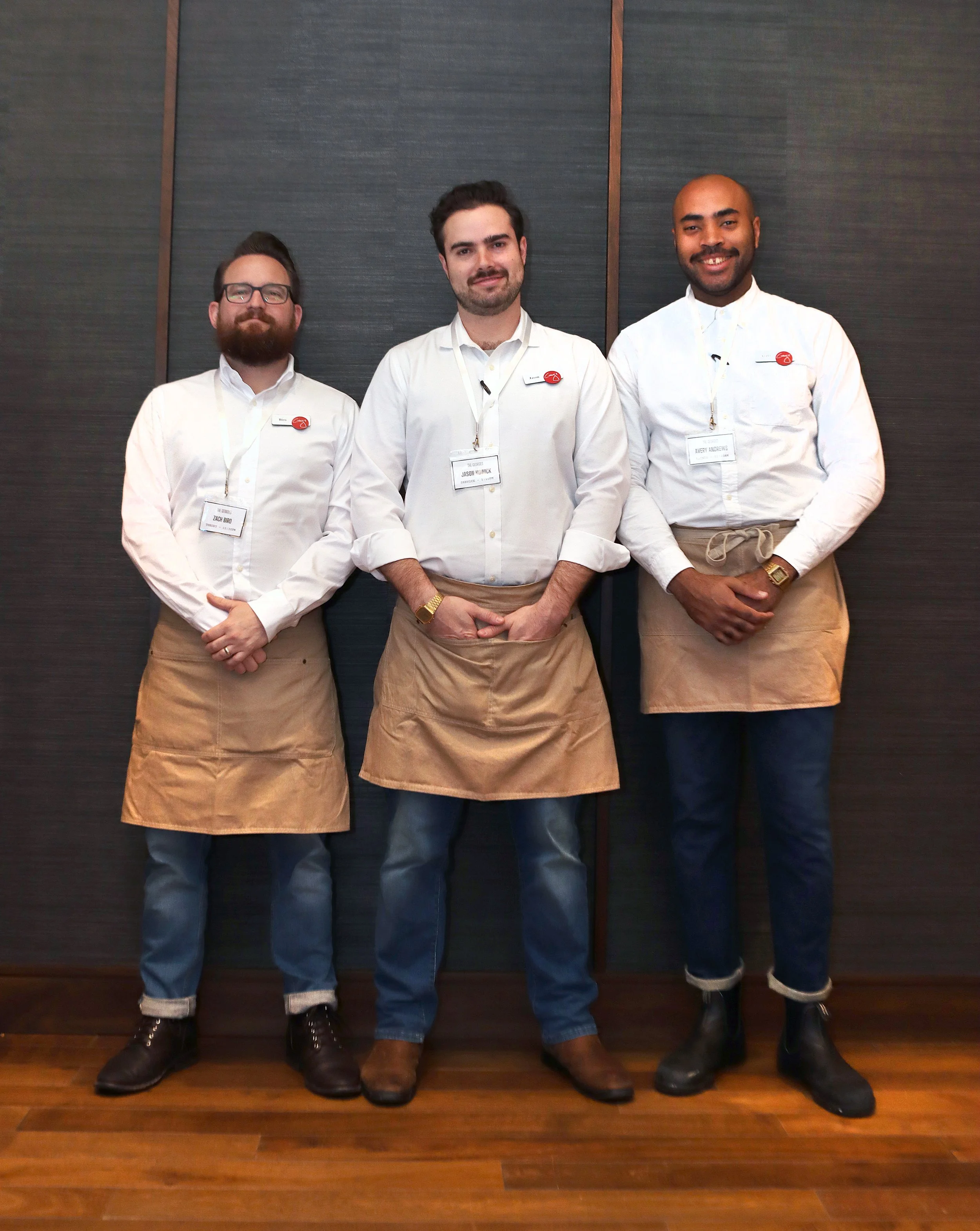 Three men standing side by side indoors against a dark wall, wearing white shirts and beige aprons with name tags. All are smiling and looking at the camera.