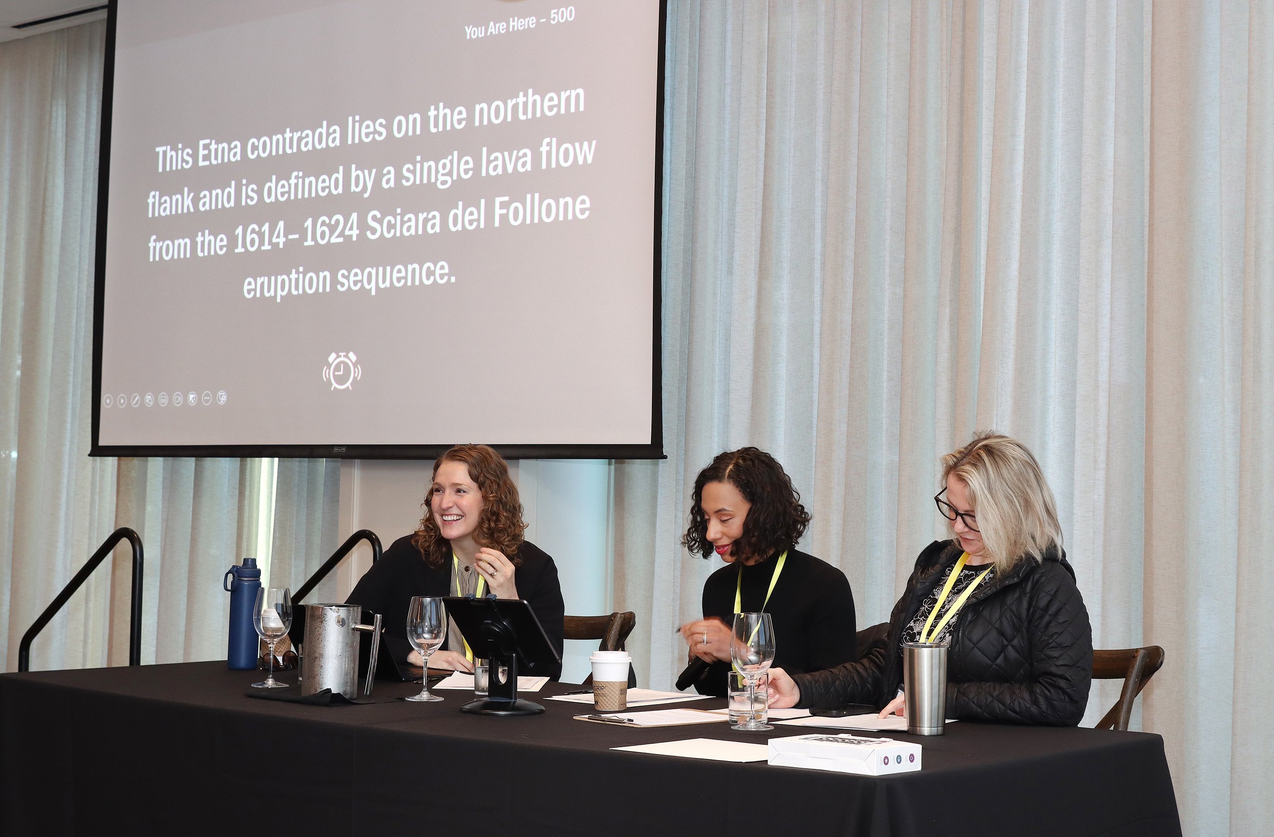 People seated at a table during a conference or panel discussion, with a large screen displaying text behind them.