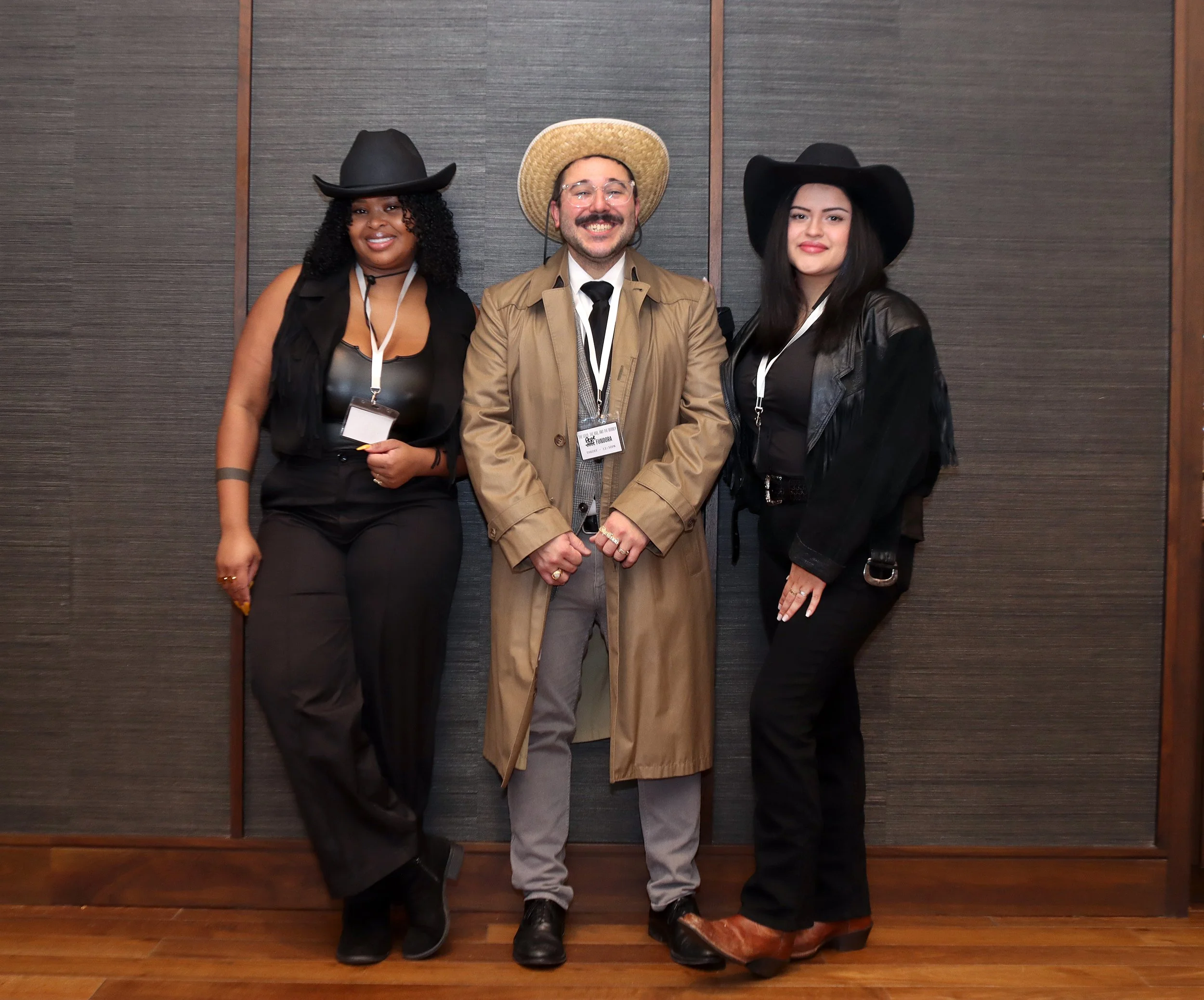 Three people dressed in cowboy-themed attire standing in front of a wooden wall. The person on the left is a woman wearing a black cowboy hat, black vest, and black pants. The person in the middle is a man with glasses, a beard, and a mustache, weari