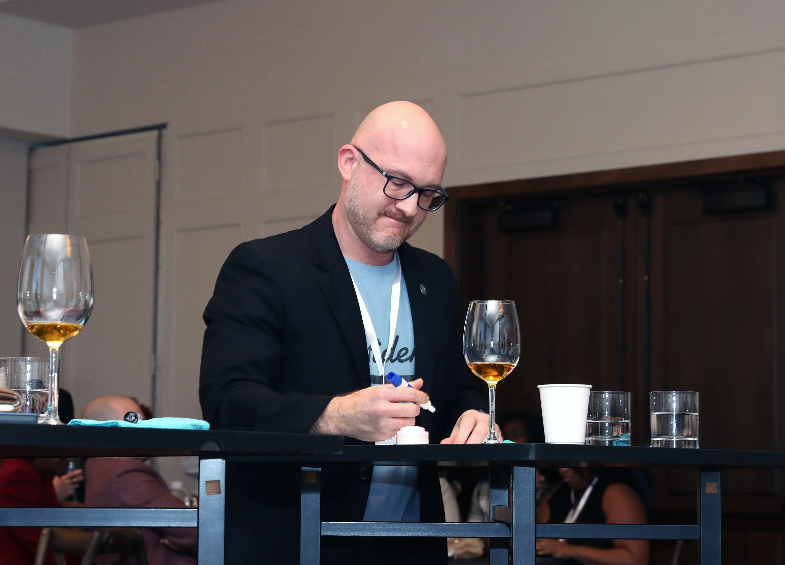 A man with glasses and a bald head is writing on a glass with a marker, in front of wine glasses and a white cup, at an indoor event with attendees in the background.