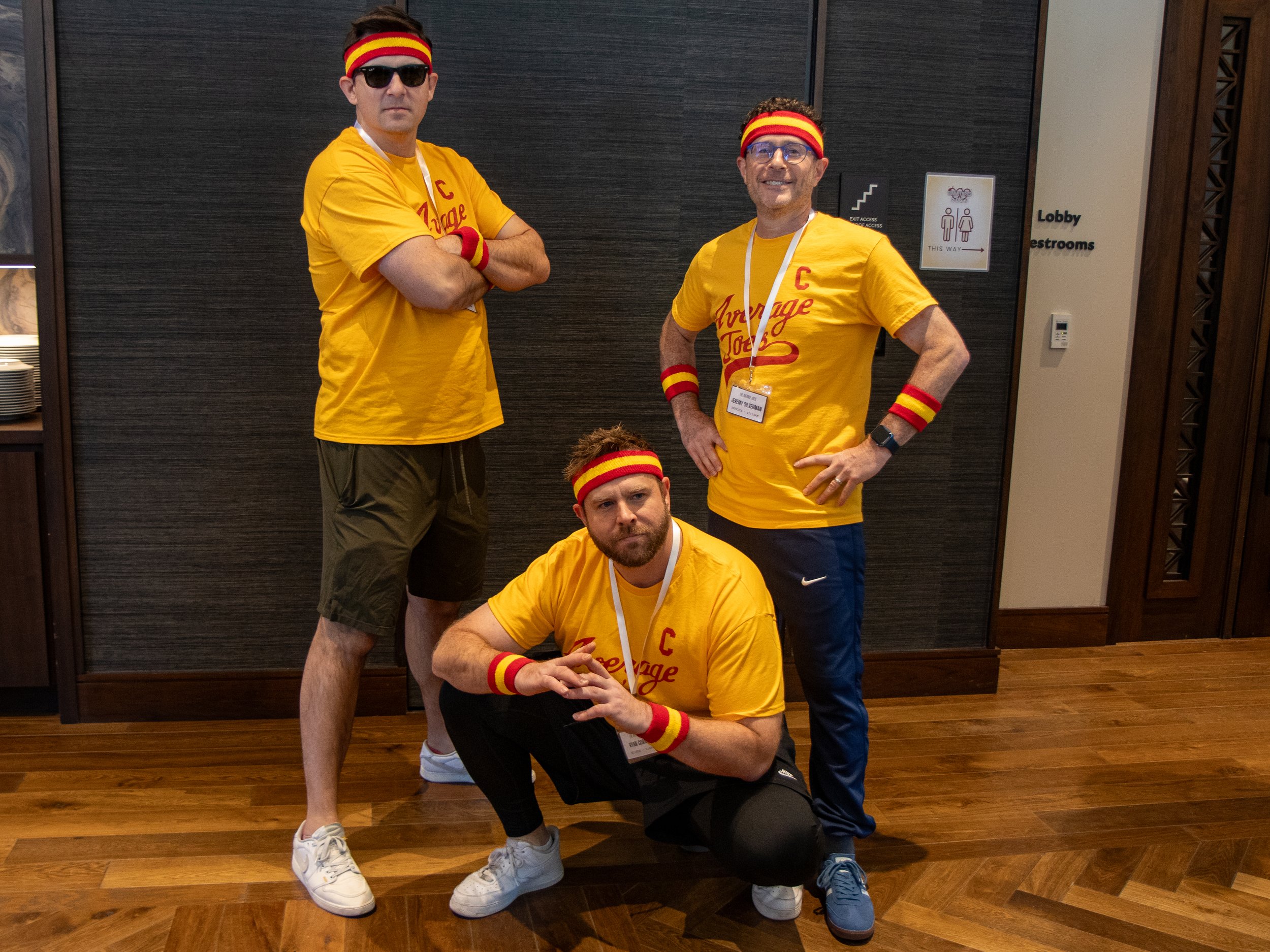 Four men in yellow T-shirts and red and yellow headbands, some with sunglasses, posing indoors in front of a dark wall. Two are standing with arms crossed, one is crouching with hands clasped, and the fourth is sitting on the floor in front.