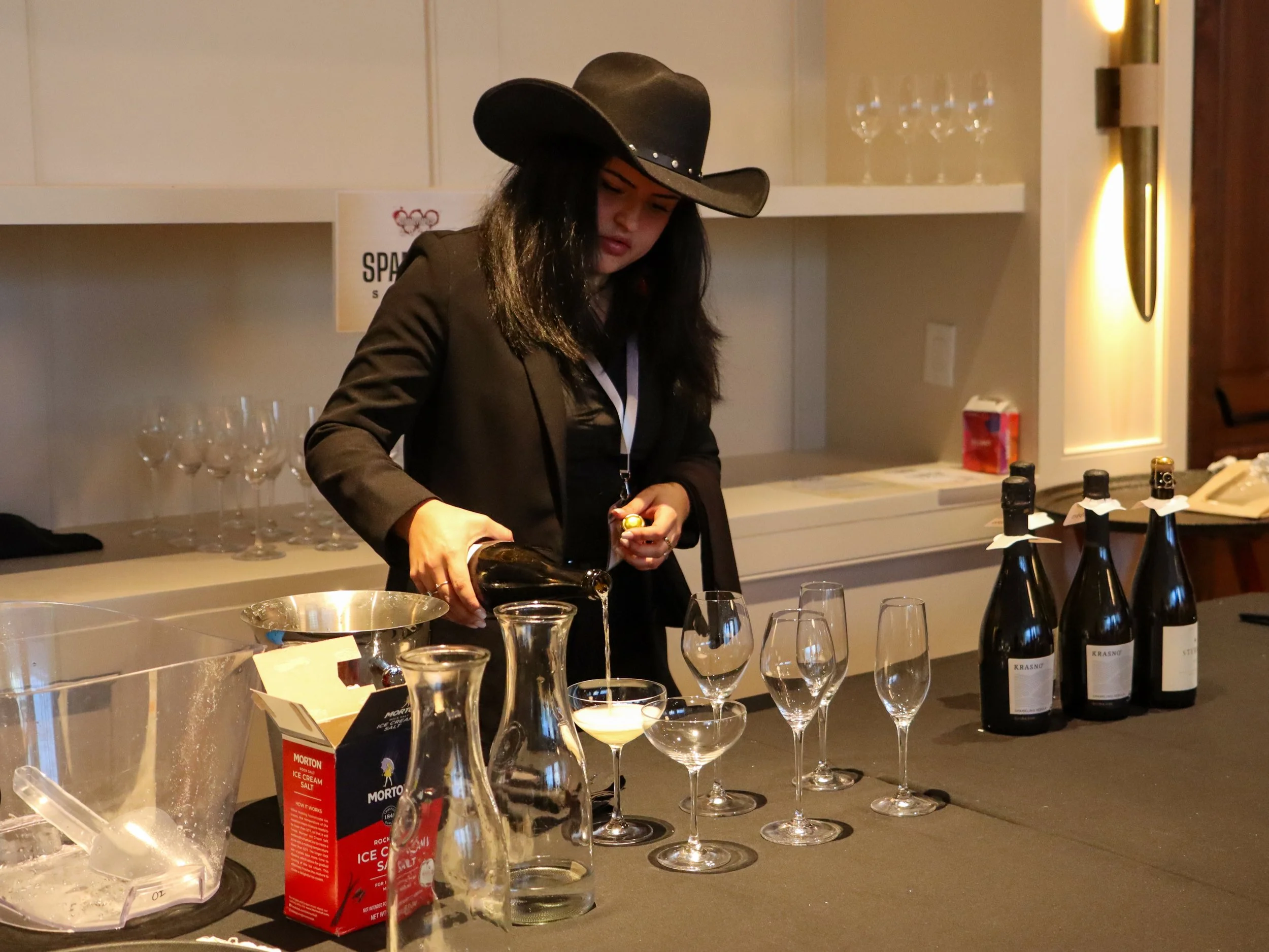 A woman in a cowboy hat pouring wine at a table with wine bottles and glassware, in a modern indoor setting.