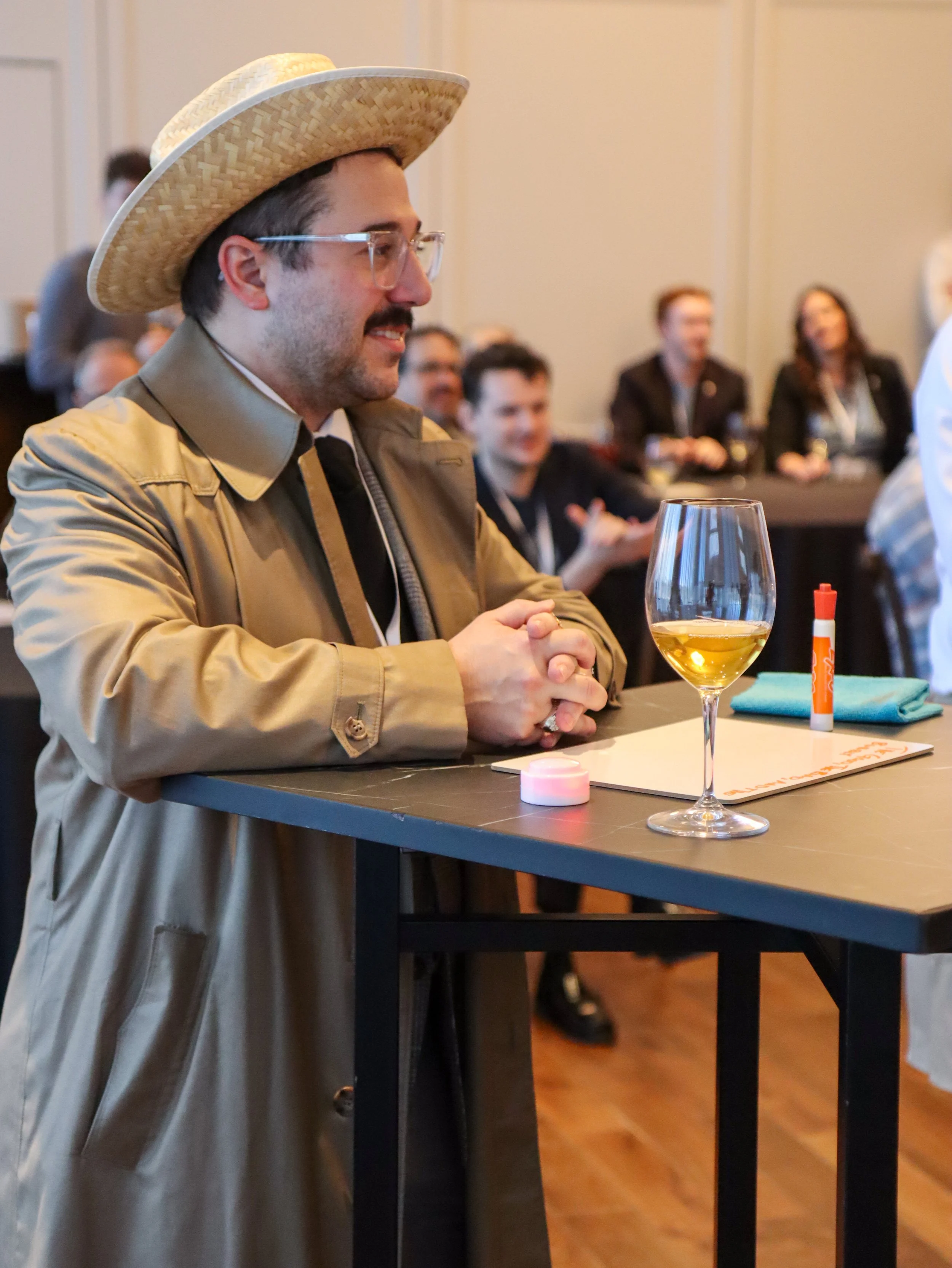 A man wearing glasses, a beige jacket, and a large straw hat sitting at a table with a glass of white wine. There are other people in the background, some smiling, at what appears to be a social or professional event.