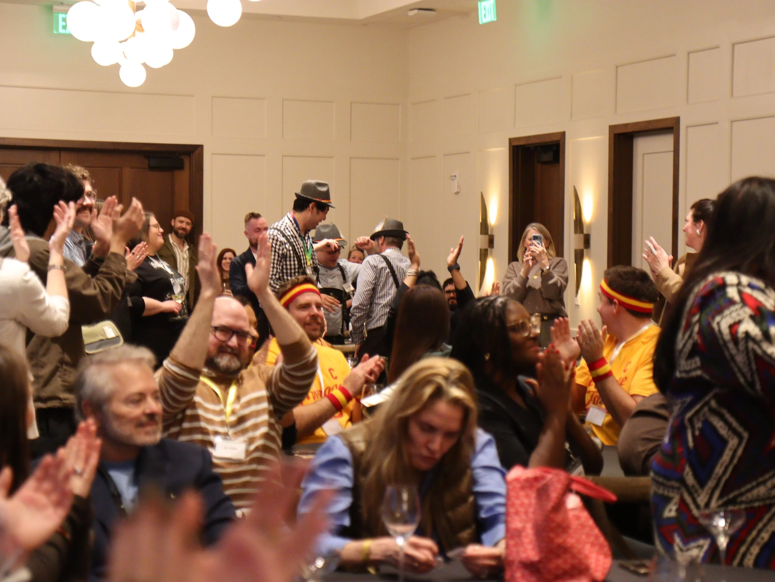 A group of people in a large room, some wearing yellow shirts and others in costumes like hats, gathered for a celebration or event, clapping and smiling.