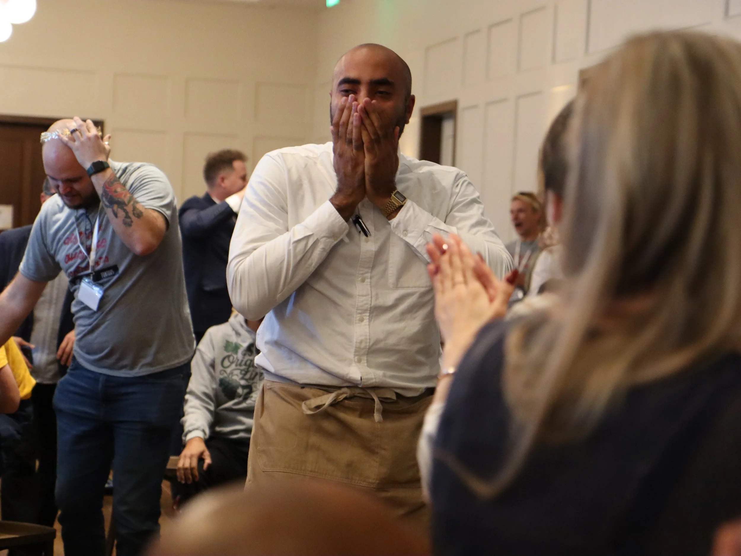 A group of people at a conference, focused on a man in a white shirt and khaki pants who appears to be surprised or emotional, with others around him visibly reacting or engaged.