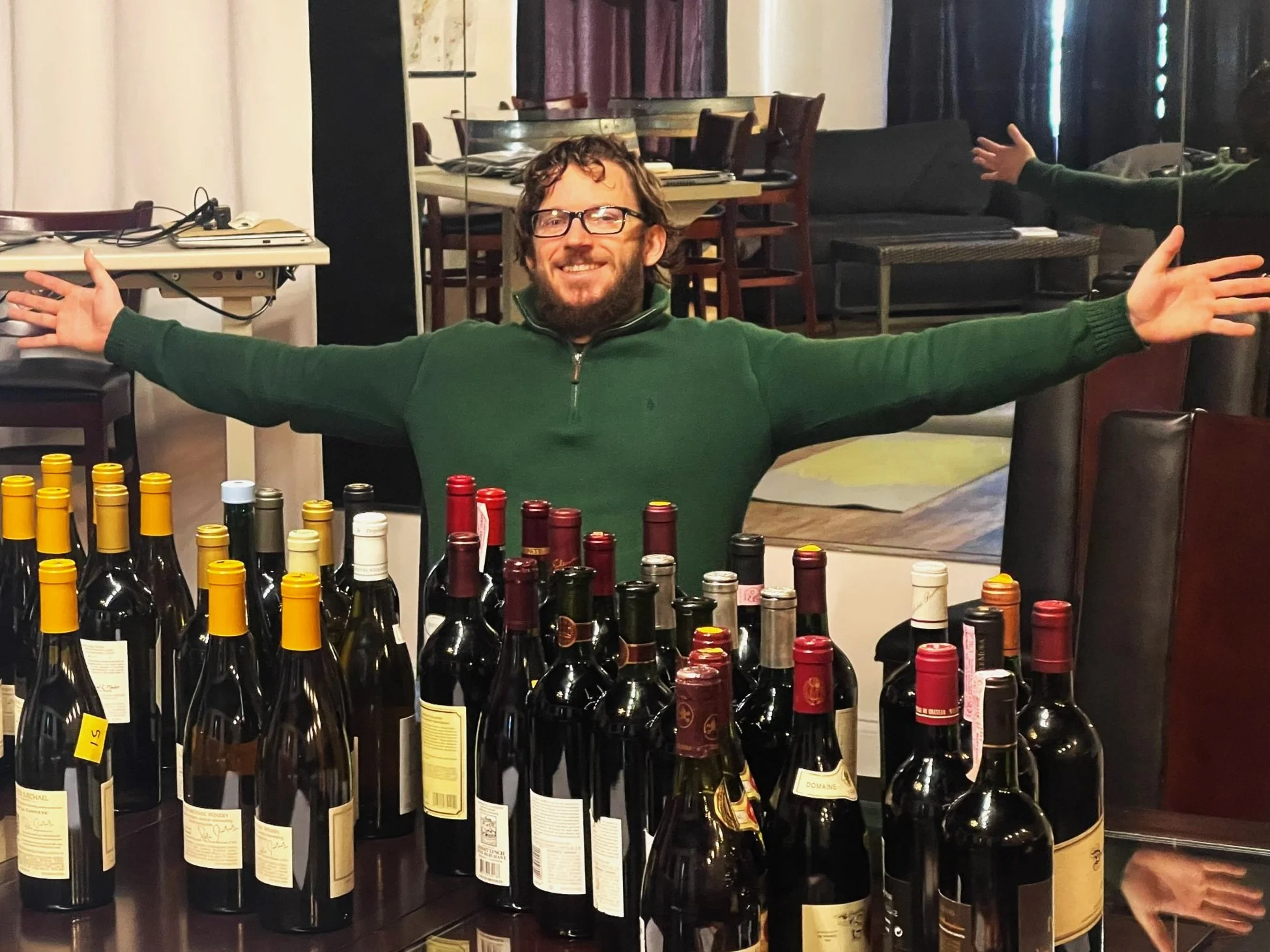 A man with glasses, a beard, and a green sweater, smiling with arms outstretched behind a table filled with wine bottles.